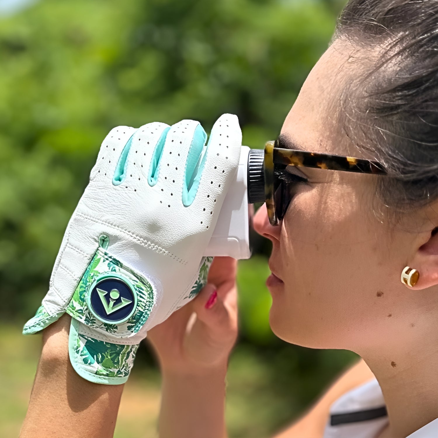 Woman golfer wearing a white tropical palm golf glove with a green VivanTee ball marker using a range finder outdoors.