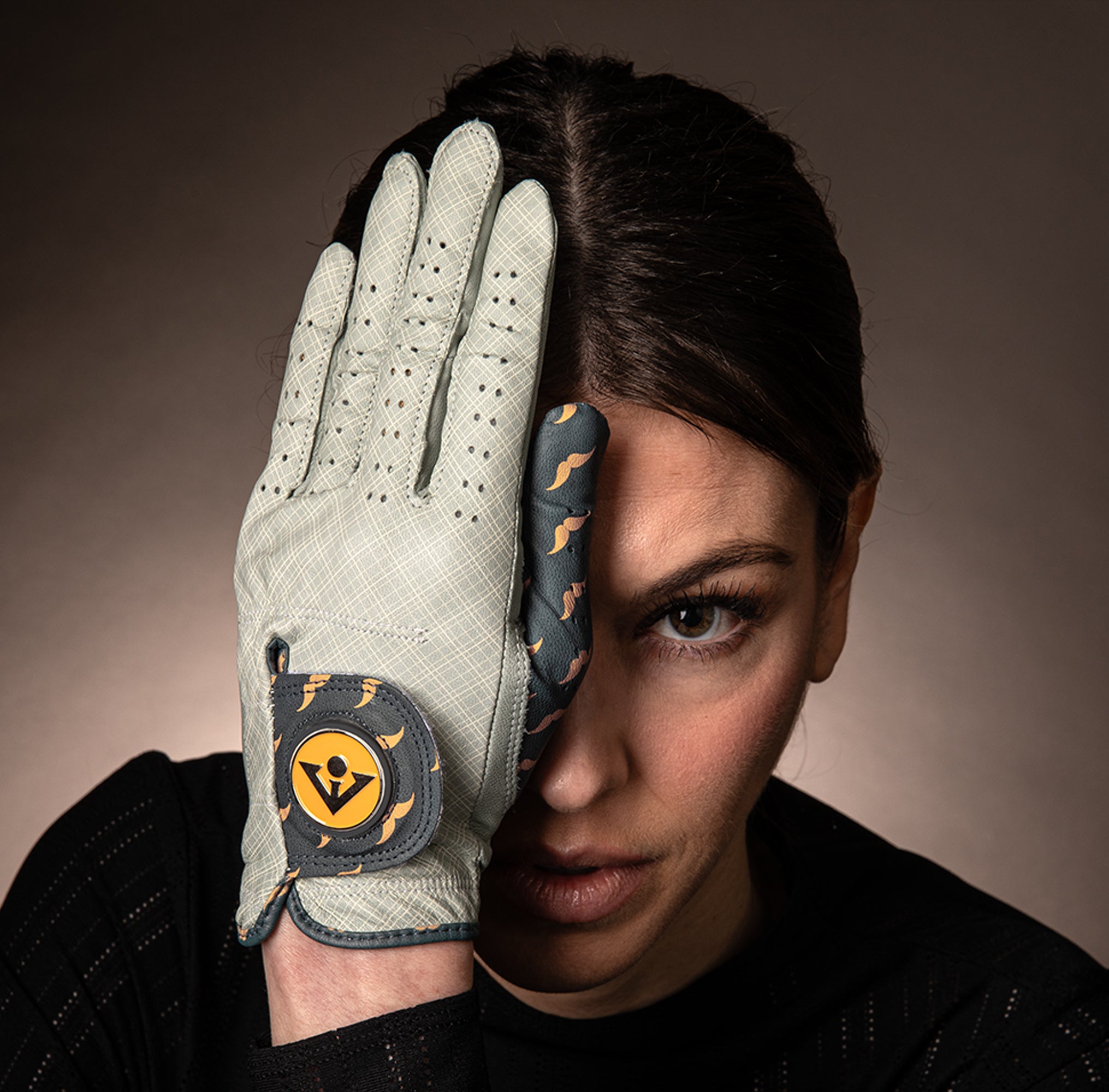 Woman wearing a mustache patterned golf glove with ball marker in grey and orange, covering her eye with a dark background.