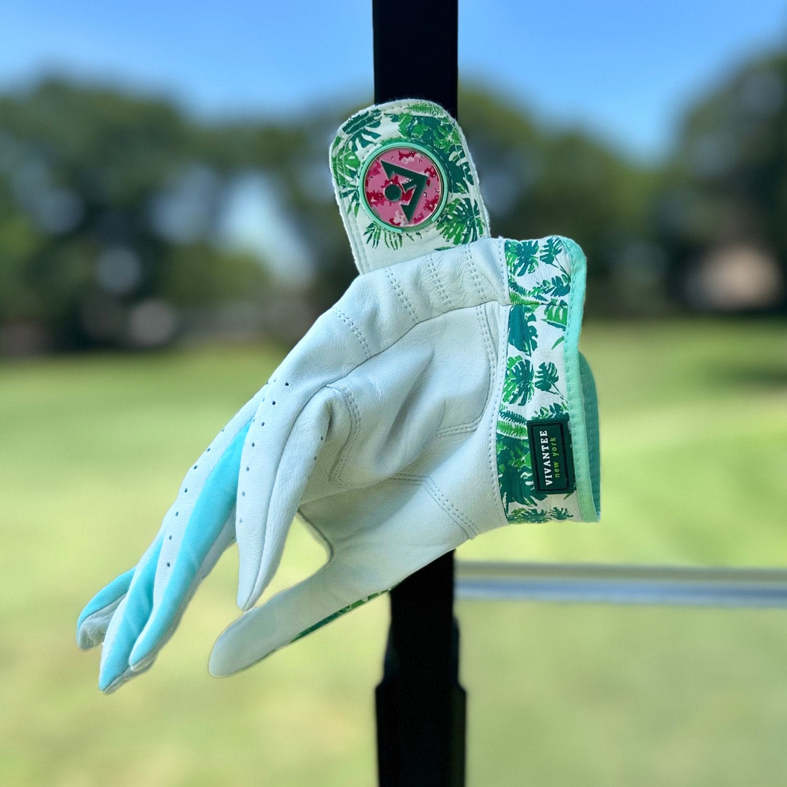 Tropical green golf glove with magnetic ball marker hanging on the post of a golf cart with a pink ball marker.