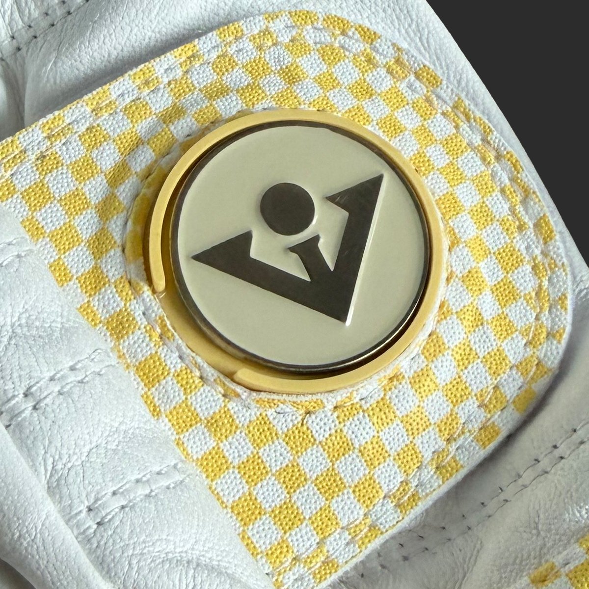 Close-up of a golf glove with a butter yellow and white checkered pattern on the back, featuring a circular gold-toned magnetic clip with a black logo.