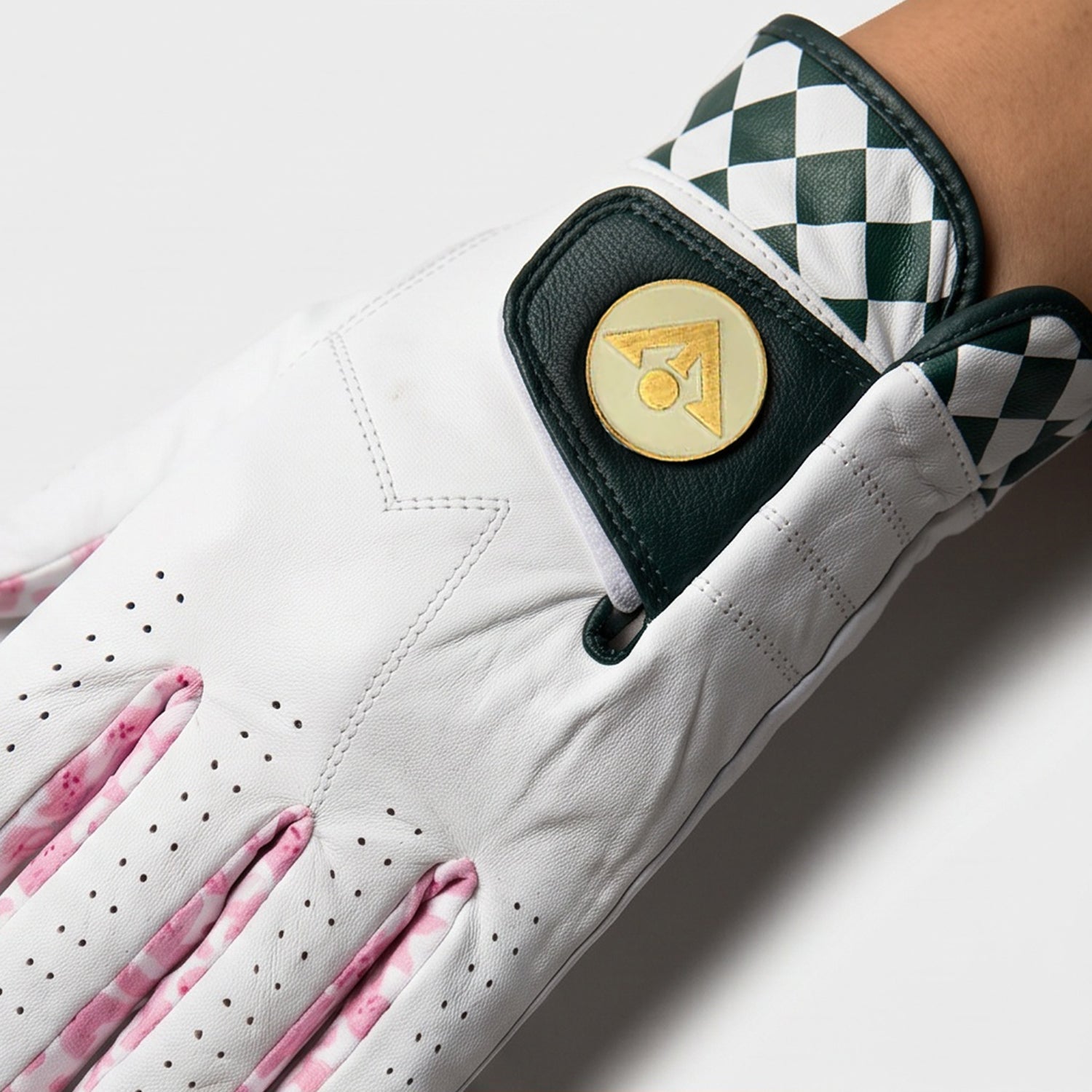 Close-up of a white golf glove with pink accents and a checkered pattern on the back, featuring a gold brand logo.