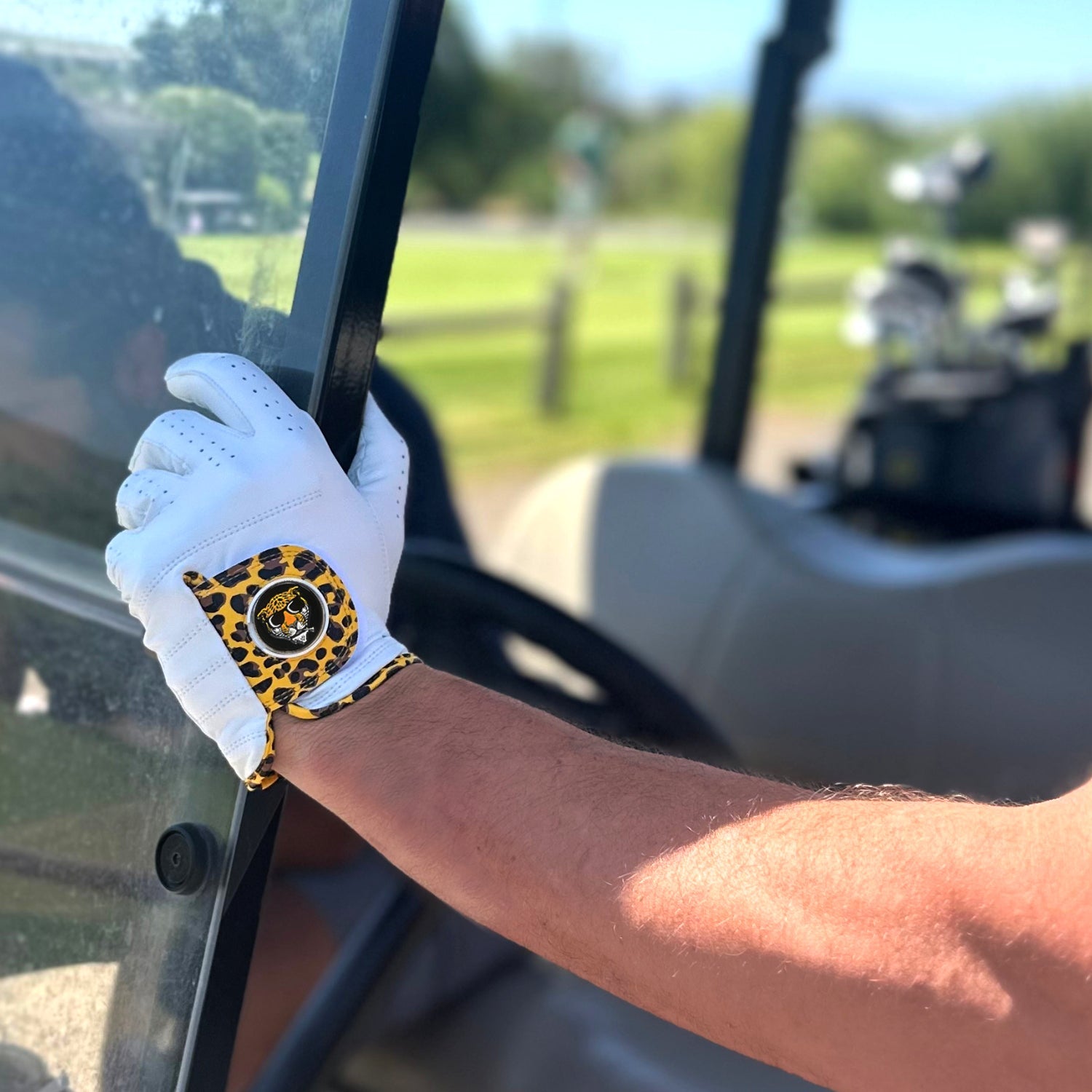 VivanTee Golf Cheetah print golf glove with magnetic ball marker grabbing the side of a Golf Cart with another golfer in the background.