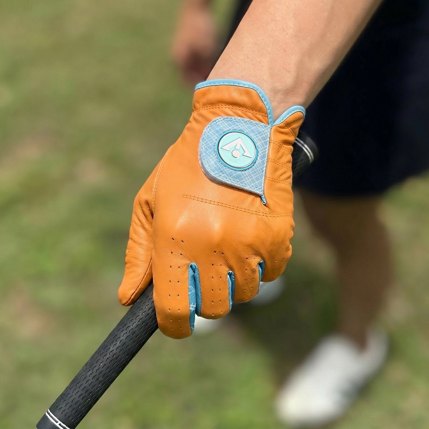 A golfer in a woman's orange golf glove holding up a club and showing the glove, with her skirt and shoes blurred in the background.