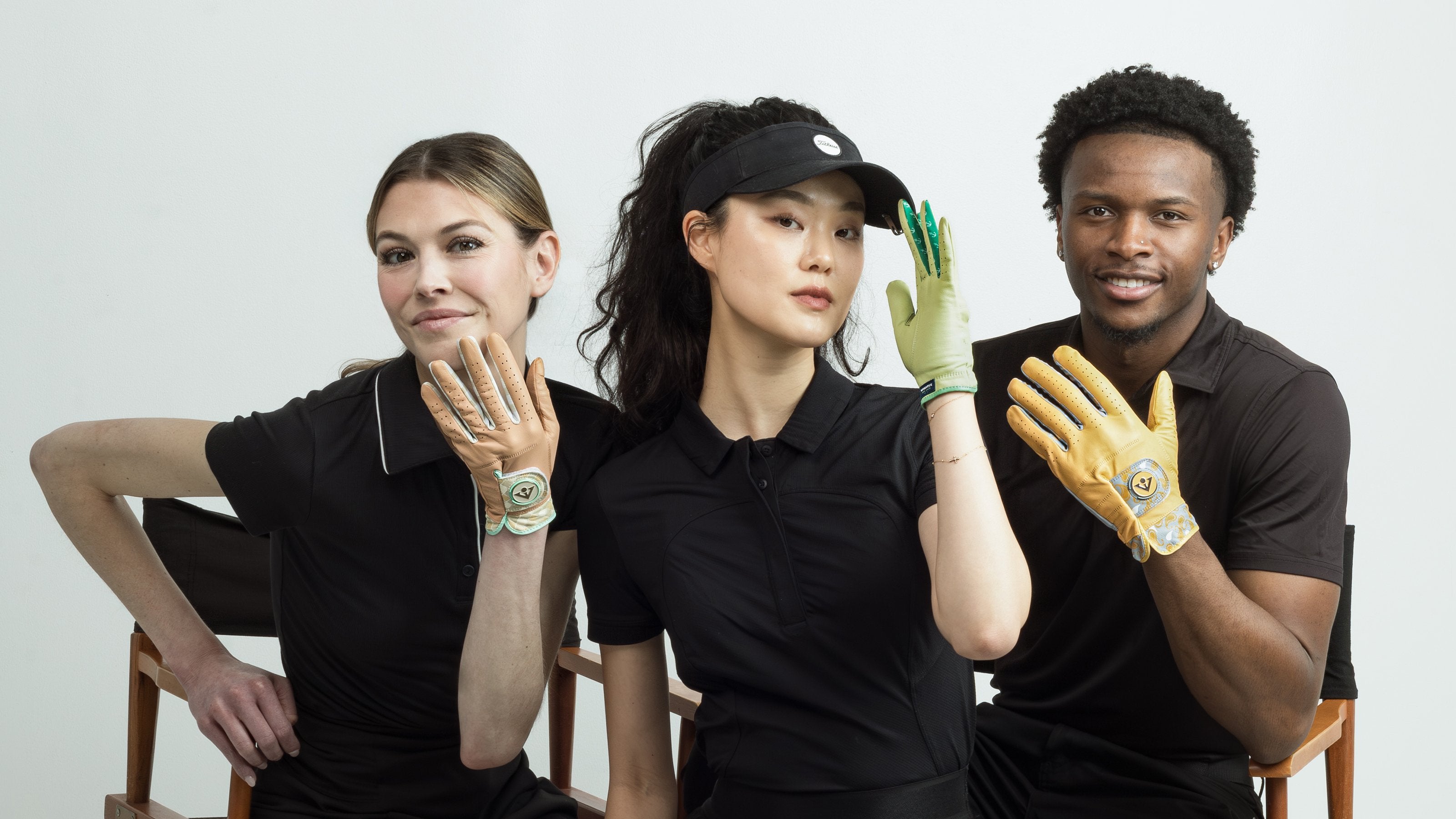 Three people wearing black outfits colorfful golf gloves with magnetic ball markers, sitting on chairs against a white background