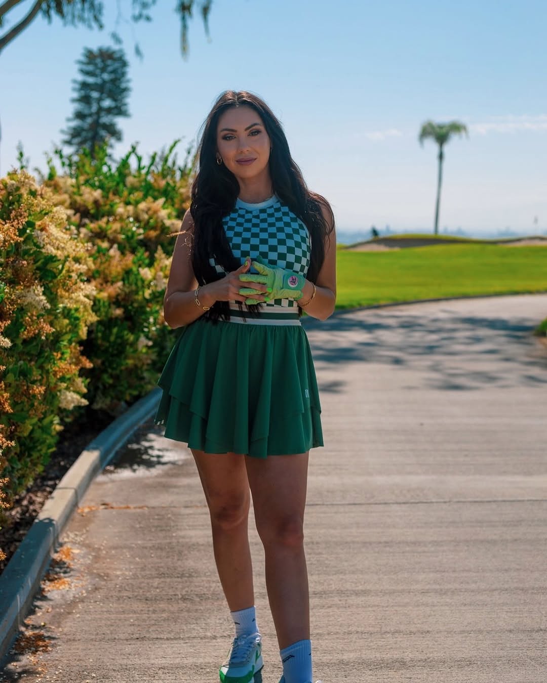 A stylish woman on a golf course wearing a checkered top and a green pleated skirt, holding a women's green golf glove with a designer logo. The golf glove features a ball marker for added convenience and style.