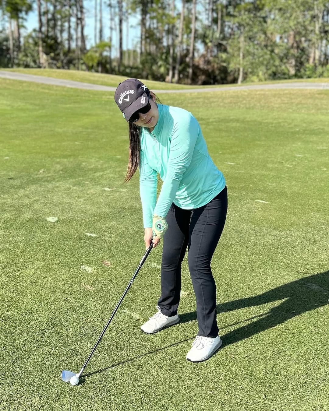 A female golfer in a light blue long-sleeve top, black golf pants, and a black Callaway cap, preparing for a shot. She is wearing a women's designer golf glove with a ball marker, which complements her elegant golf outfit.