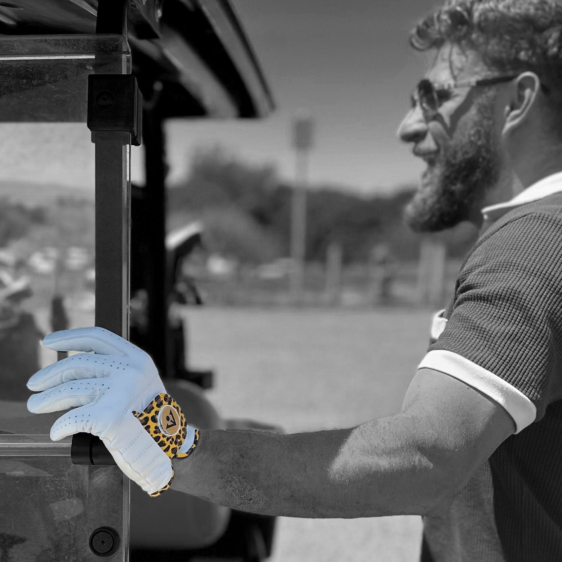 Man holding a golf cart wearing a white cheetah print golf glove with a ball marker, standing next to a golf cart on a golf course.