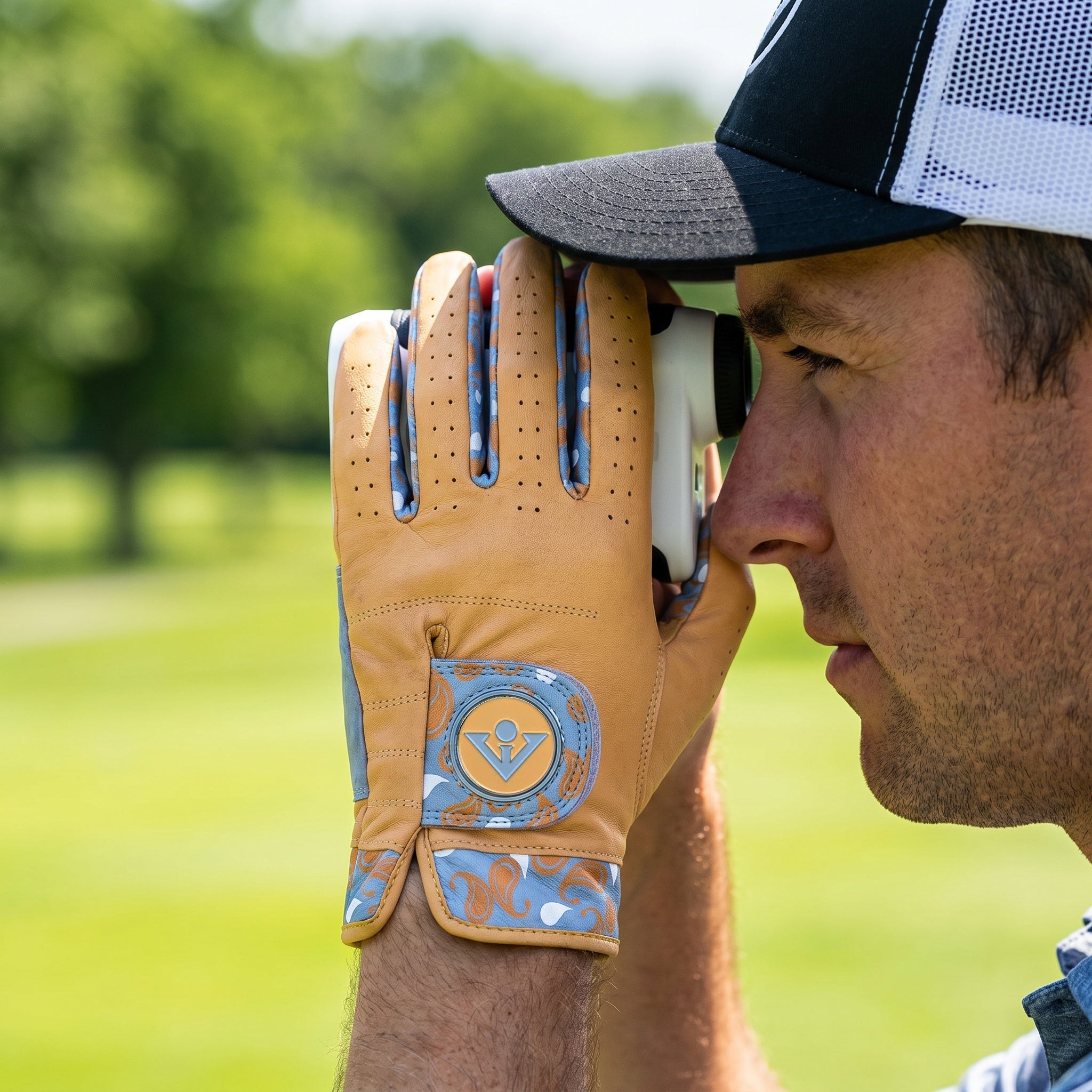 Man holding a range finder with a gold golf glove on his hand, wearing a cap, against a blurred green background.