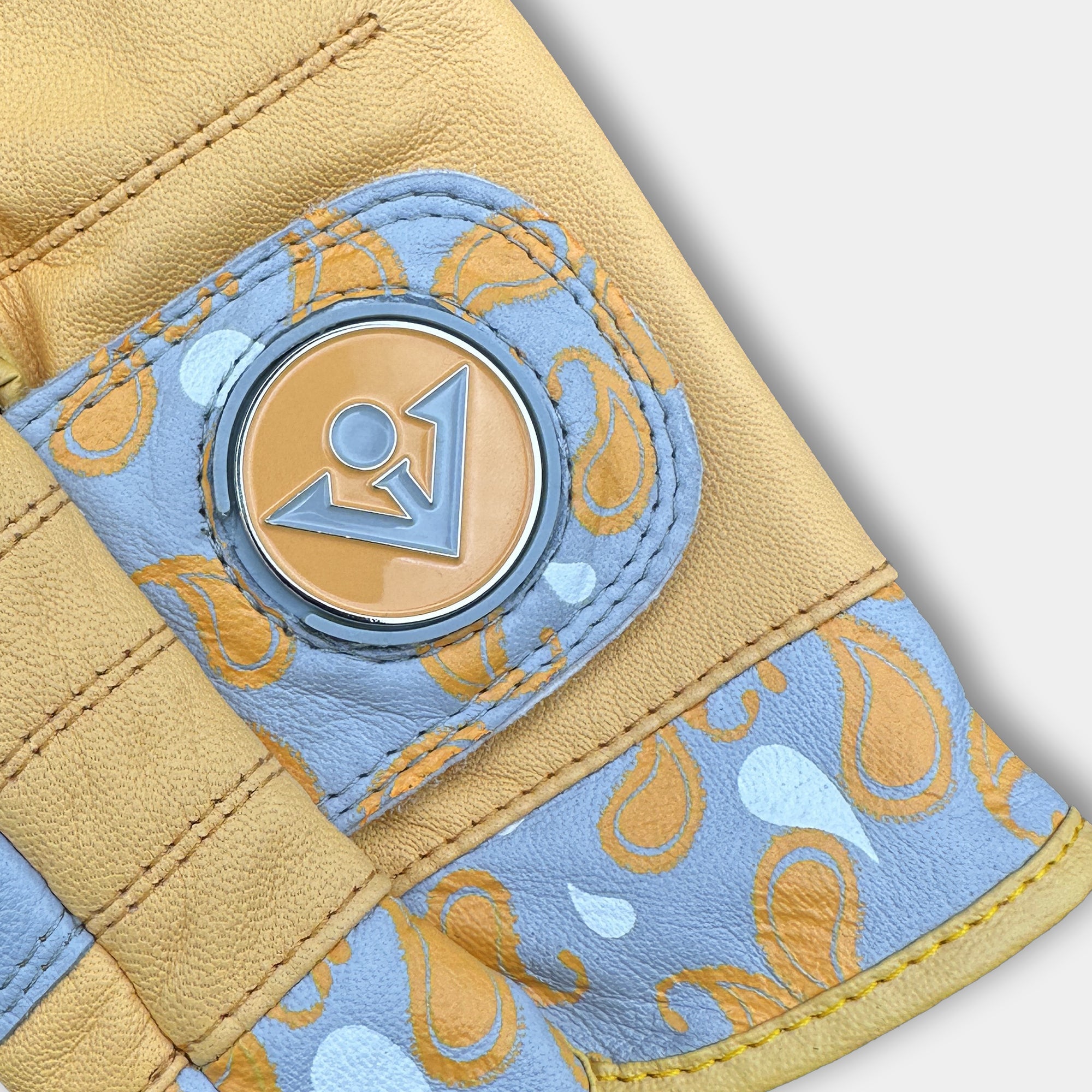 Close up of a gold golf glove with paisleys and a magnetic ball marker on a light grey background.