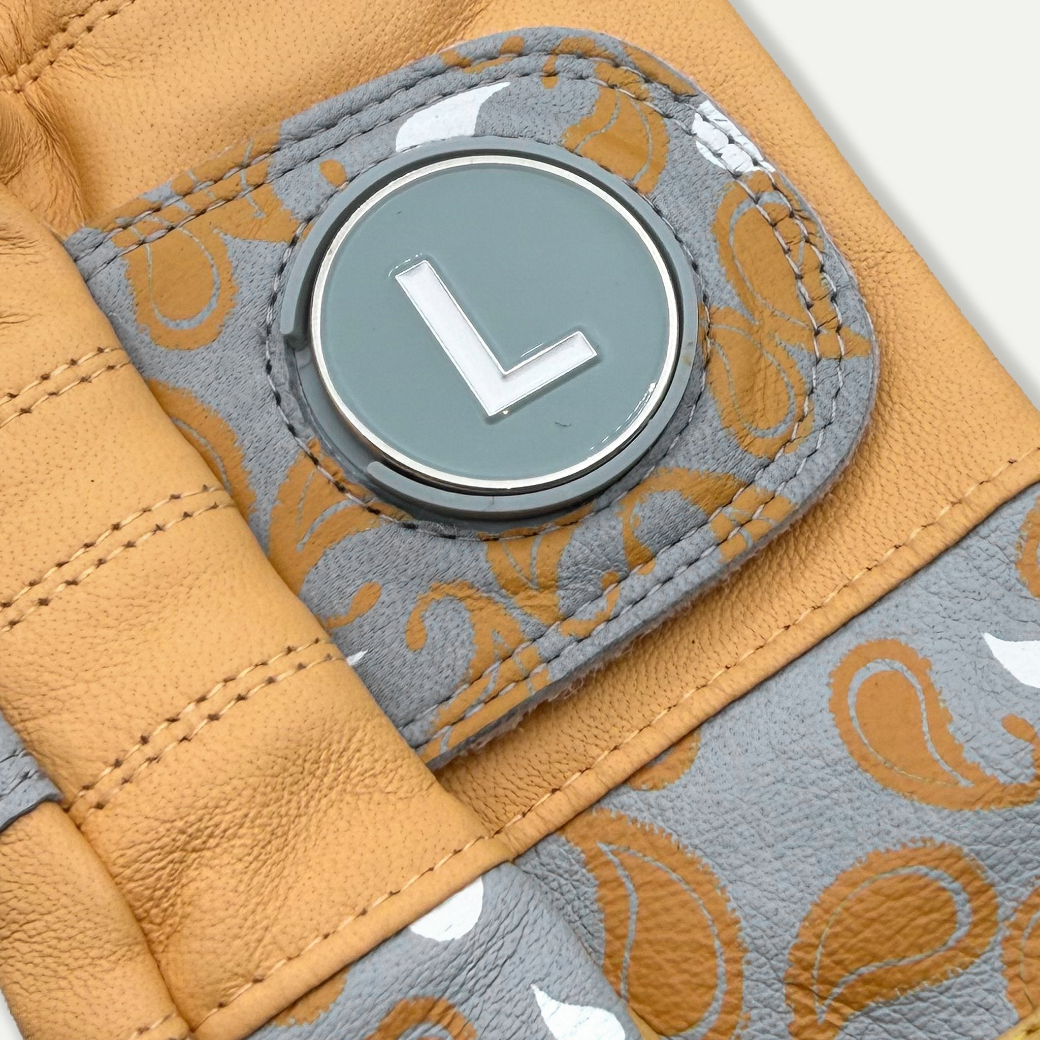 Close up of a gold golf glove with paisleys and a magnetic ball marker on a light grey background.