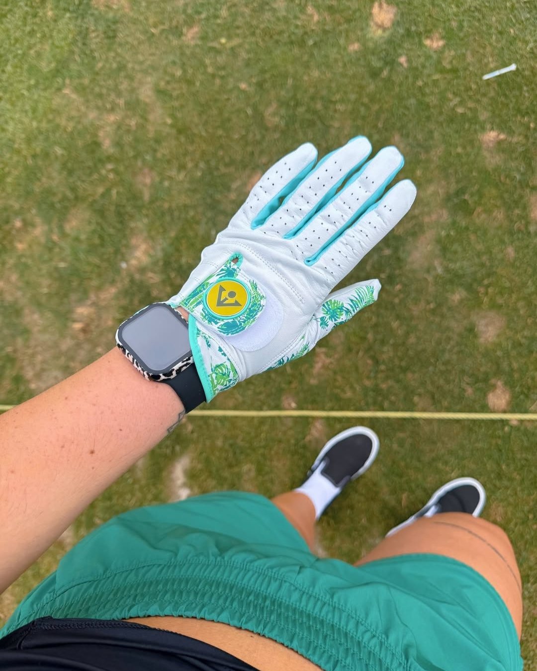 Golf glove on a person's hand with a green and white design, standing on grass.