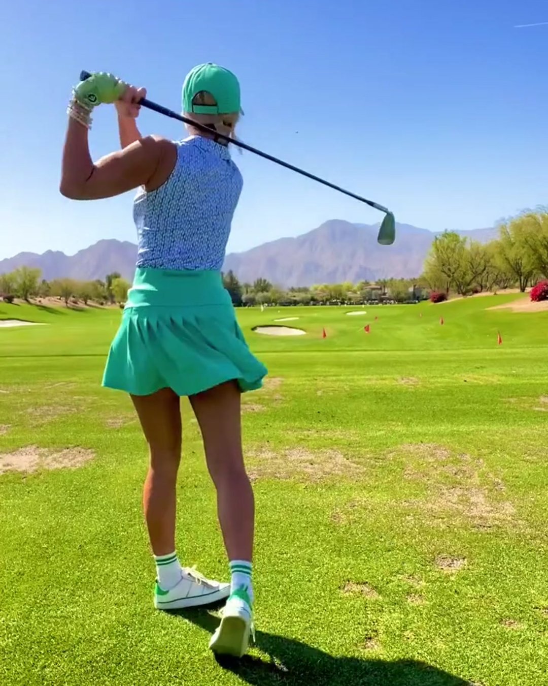 A female golfer mid-swing on a scenic golf course with mountain views, dressed in a mint green skirt, matching cap, and a blue-patterned sleeveless top. She is wearing a women's green golf glove, adding a chic touch to her sporty ensemble.