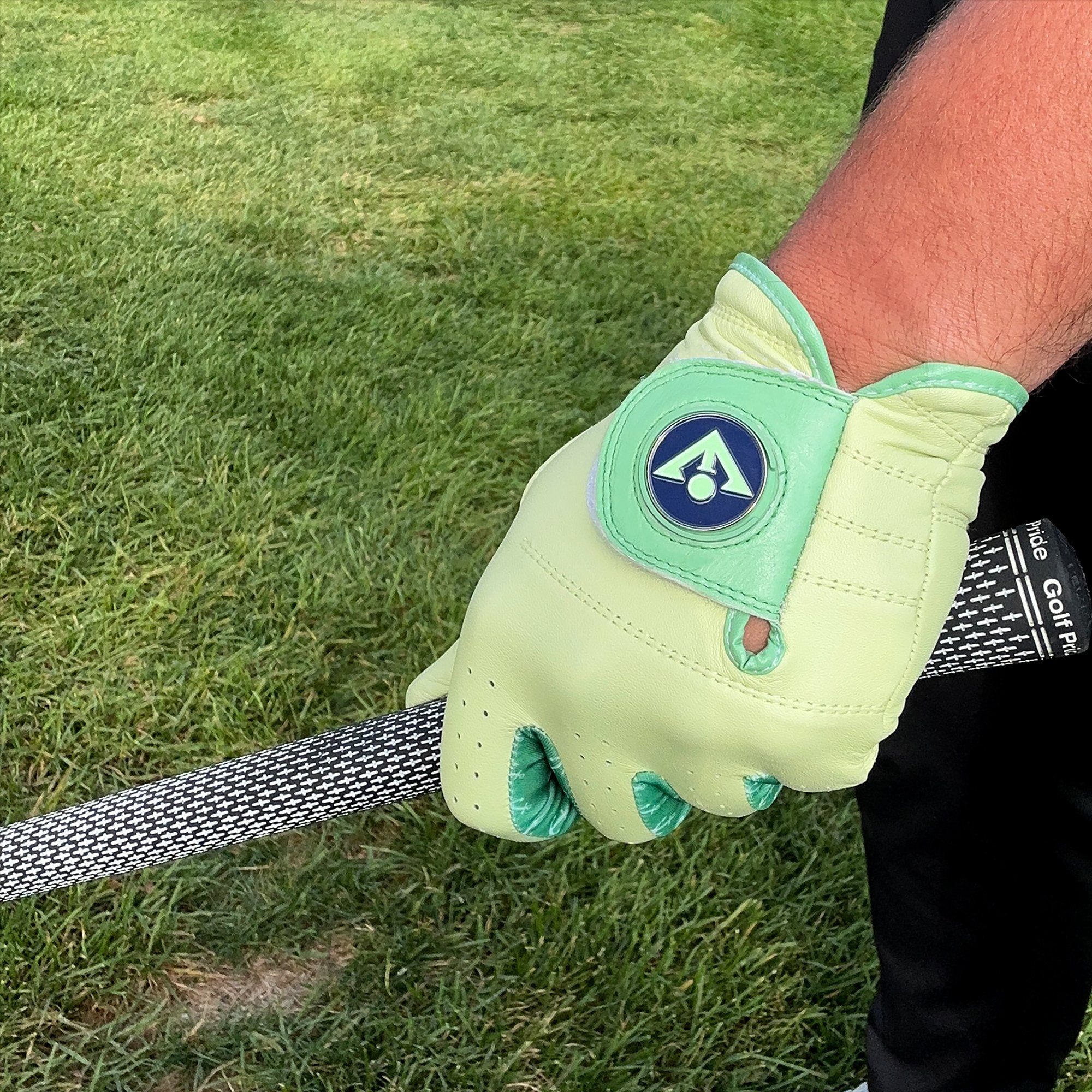 Close up of a green golf glove with blue magnetic ball marker by VivanTee golf, gripping a golf club with grass in the background.