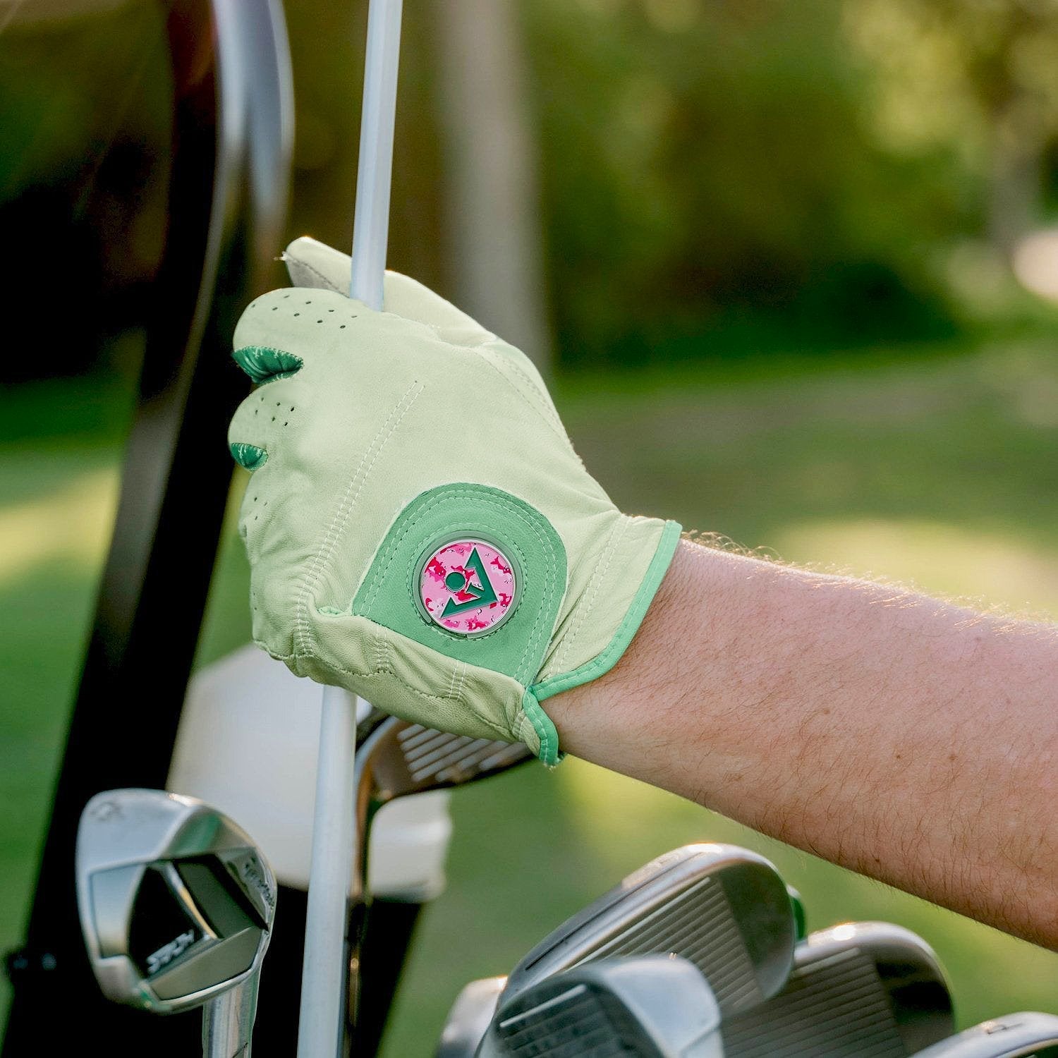 Close up of men's mint green golf glove with pink ball marker putting away a golf club in his bag.