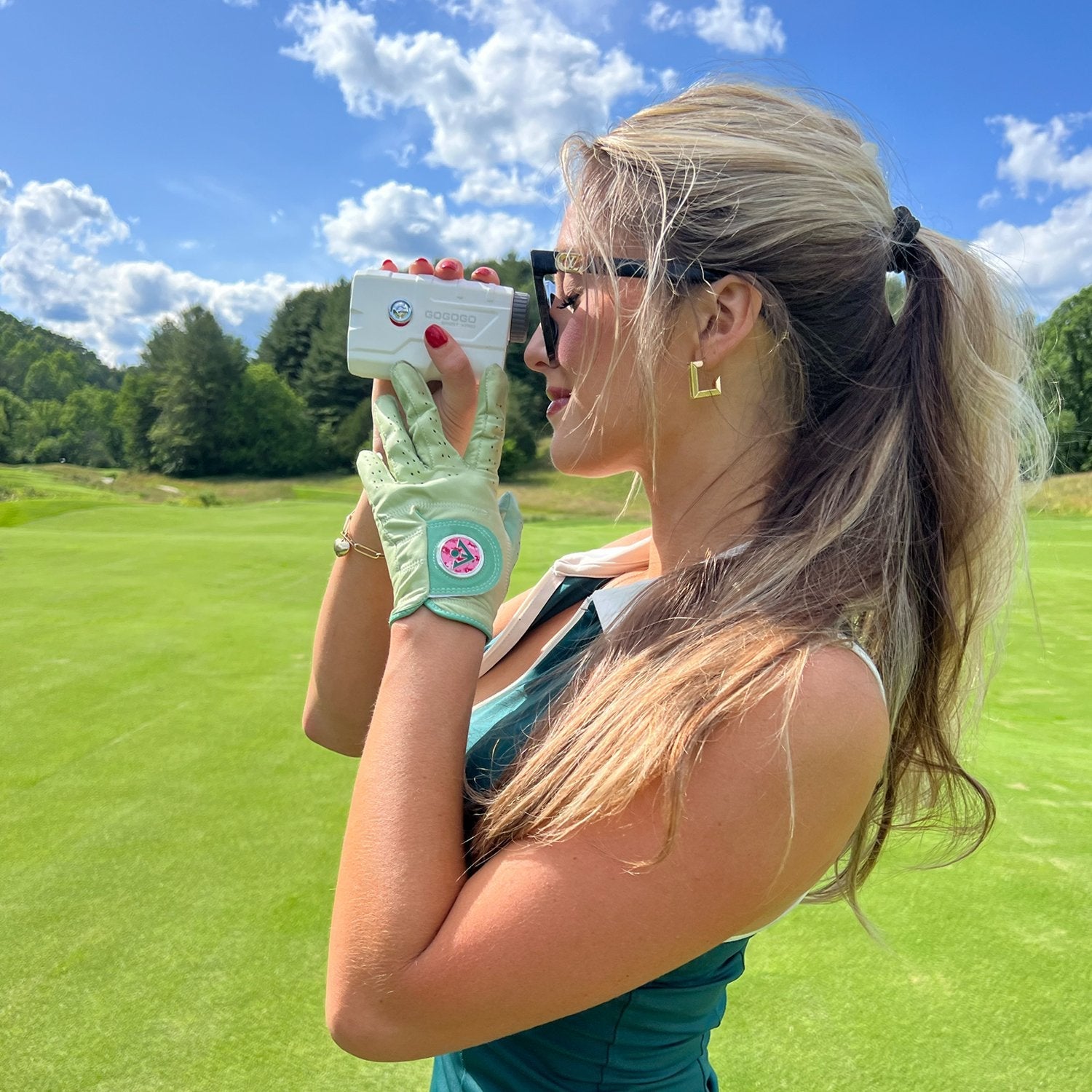 Woman in green golf glove and green dress looking at her rangefinder, golf glove sporting a pink magnetic ball marker.