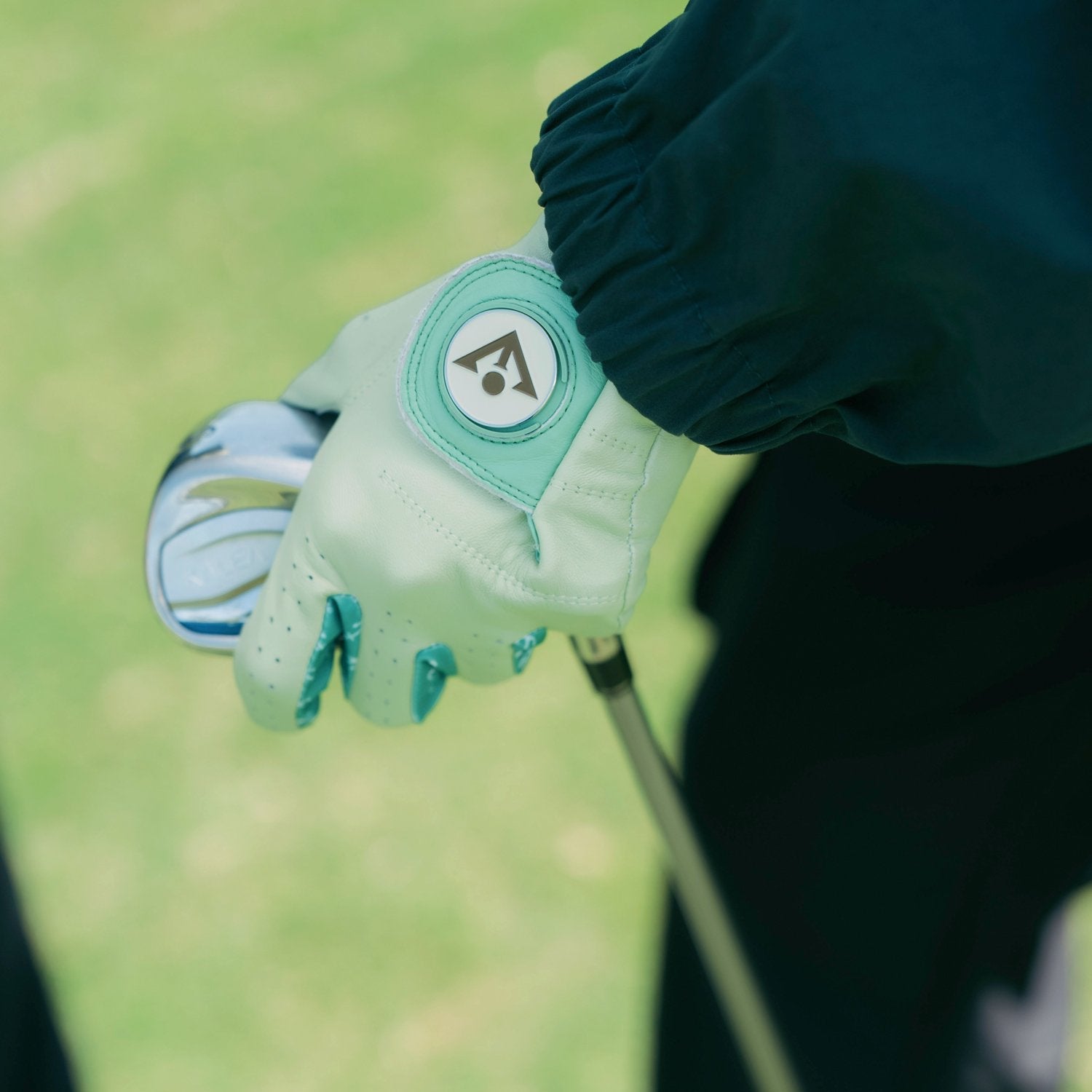 Close-up of a person wearing a light green golf glove with a VivanTee logo ball marker on a blurred background.