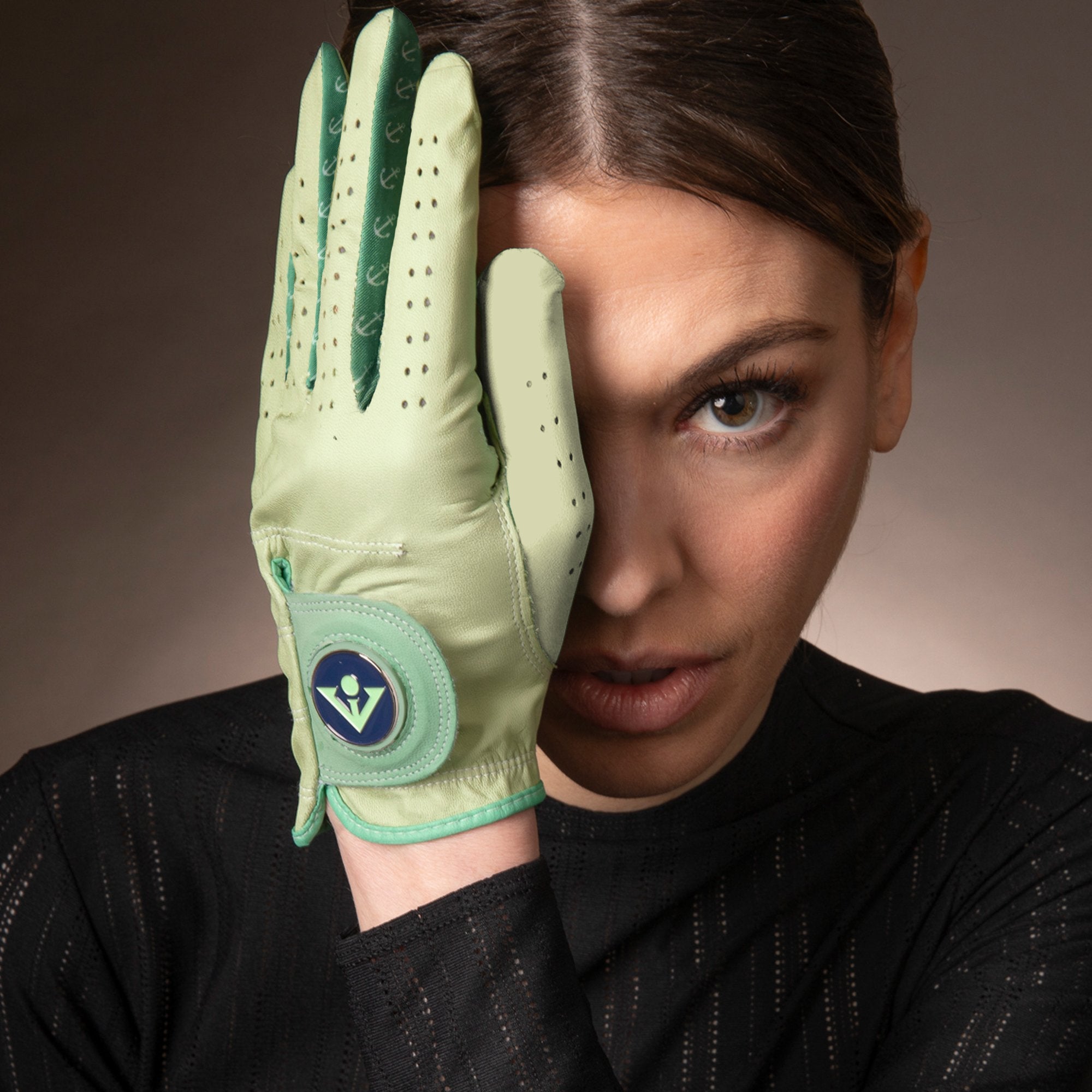 Intense gaze of a woman partially covering her face with a hand wearing a women's mint green golf glove, detailed with perforations and featuring a vibrant blue and green magnetic ball marker with VivanTee Logo..