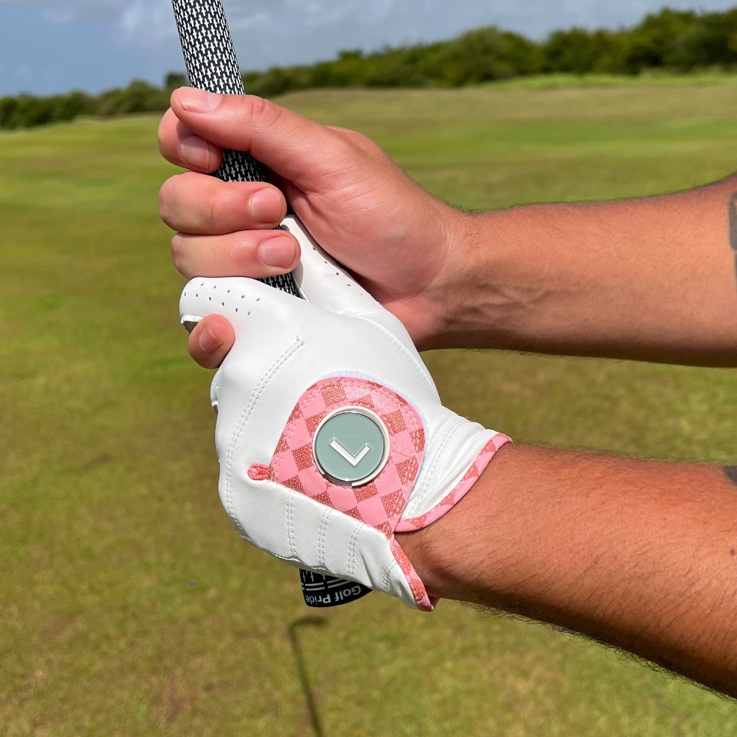 Hand wearing a white golf glove with pink accents holding a golf club on a golf course.