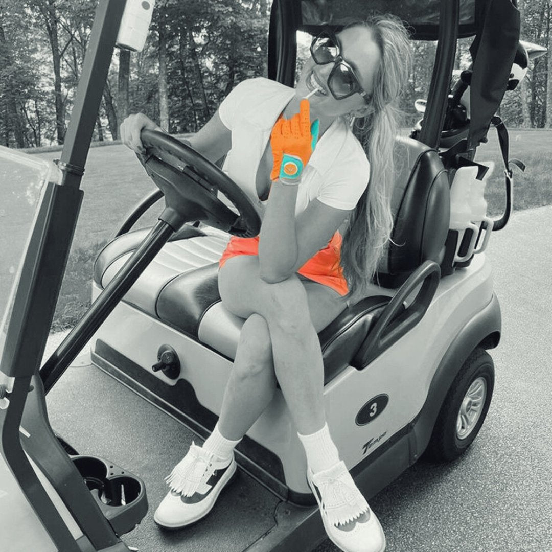 Golfer in cart wearing an orange and blue golf glove with ball marker and coral skirt, in a black and white setting, showing the colors in the skirt and glove only.