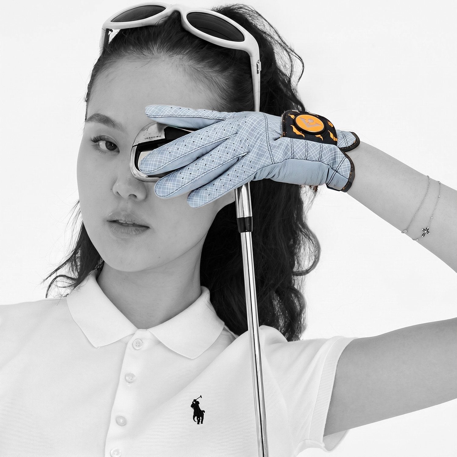 Woman wearing a white polo shirt with a logo, holding sunglasses and a golf club, with a focus on the grey golf glove.