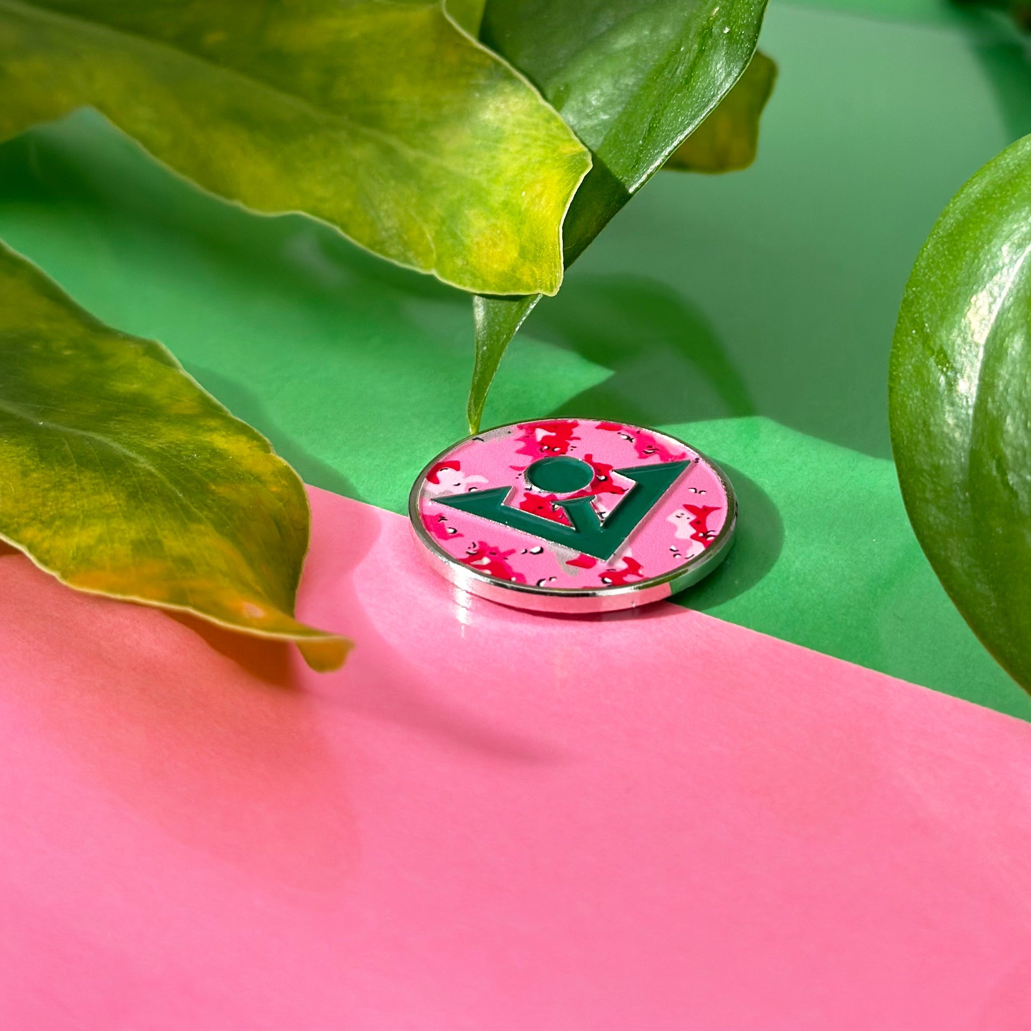 Pink camo magnetic golf ball marker, surrounded by leaves and pink and green background.