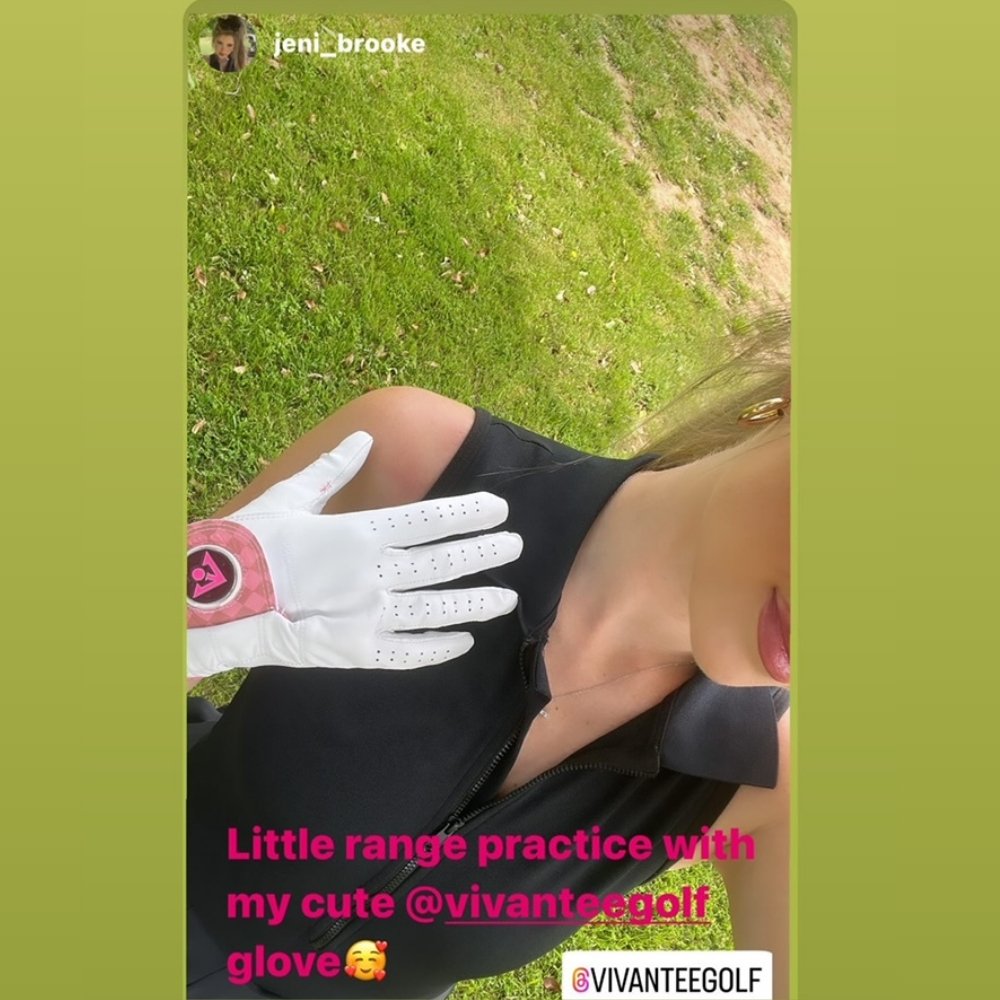 Close-up of a golfer's hand wearing a unique white golf glove with a pink magnetic ball marker on the wrist, grass background.