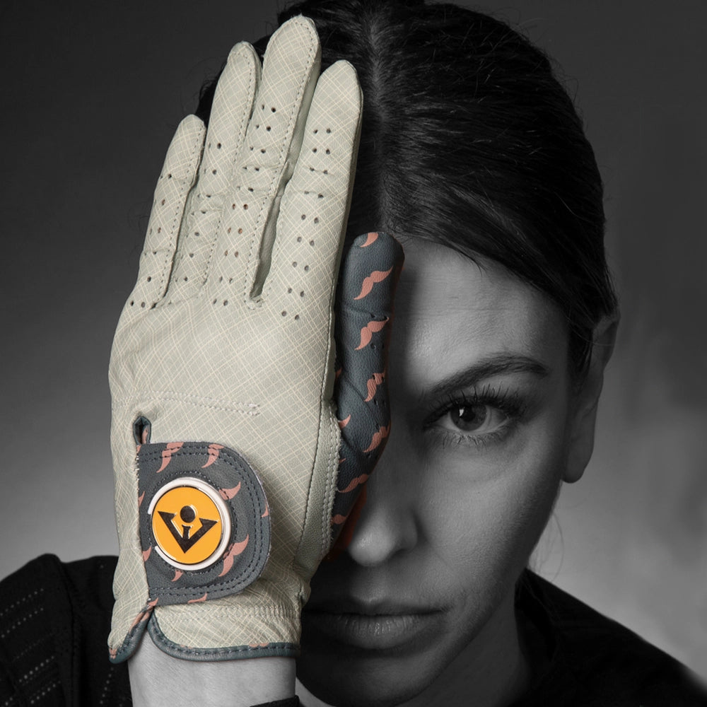 Golf glove in grey and charcoal with ball marker held in front of a female models face in a monochromatic setting.