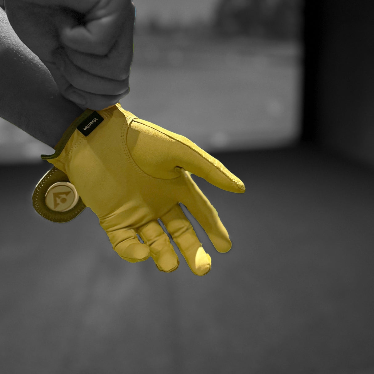 Yellow VivanTee golf glove being donned by a golfer with a golf simulator in the background, in black and white except for the golf glove.