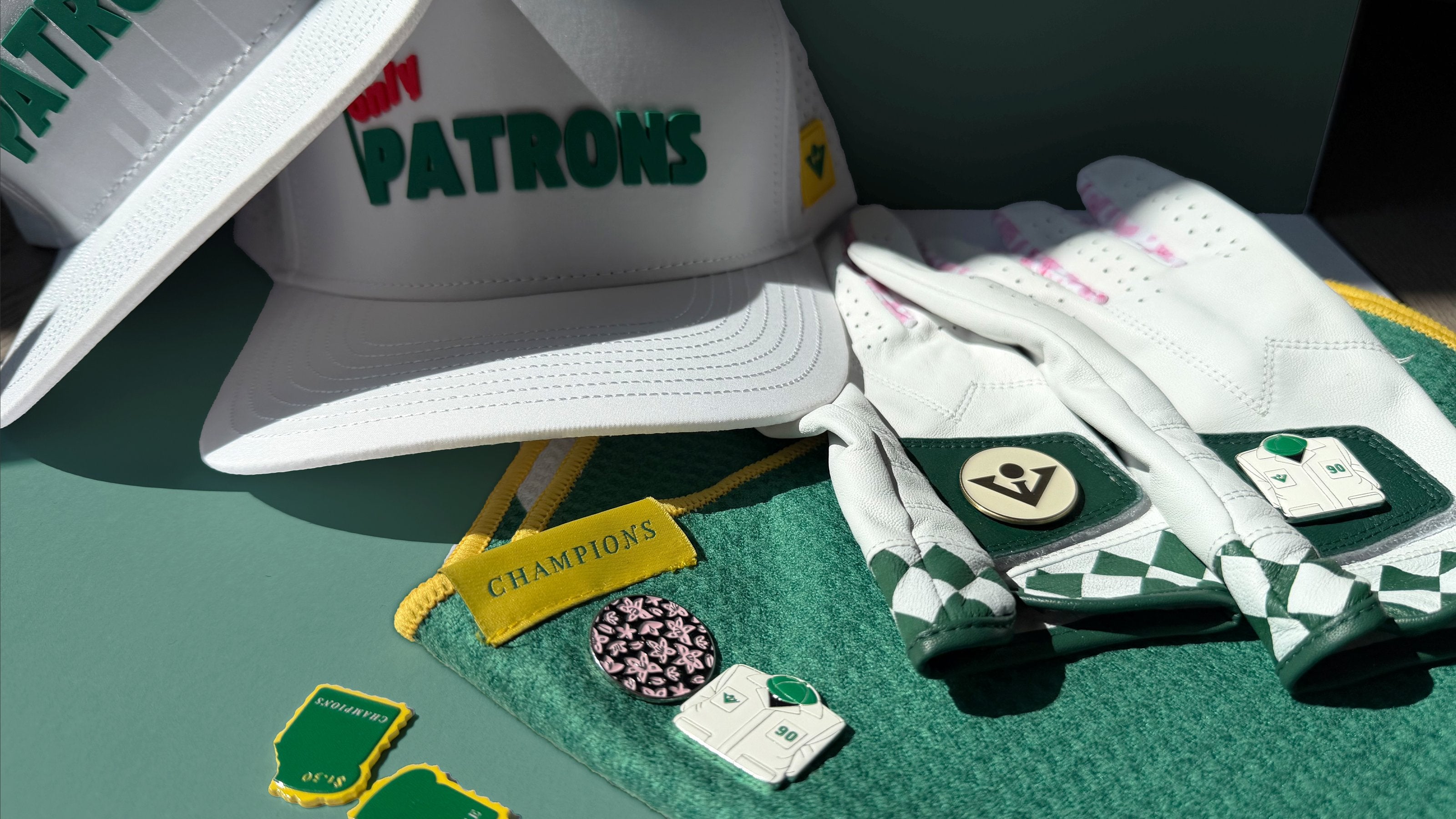 Masters inspired golf accessories, showing a White cap with 'Only Patrons' branding, white golf gloves with magnetic ball marker, and green and white caddie ball markers with a green background..