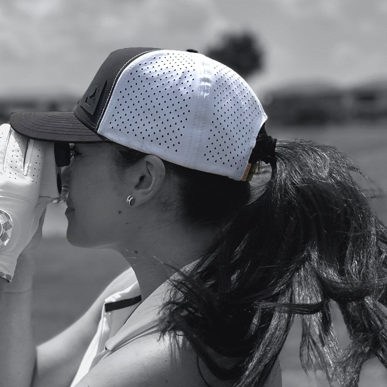 Woman wearing a golf performance hat and golf glove using a range finder in black and white.