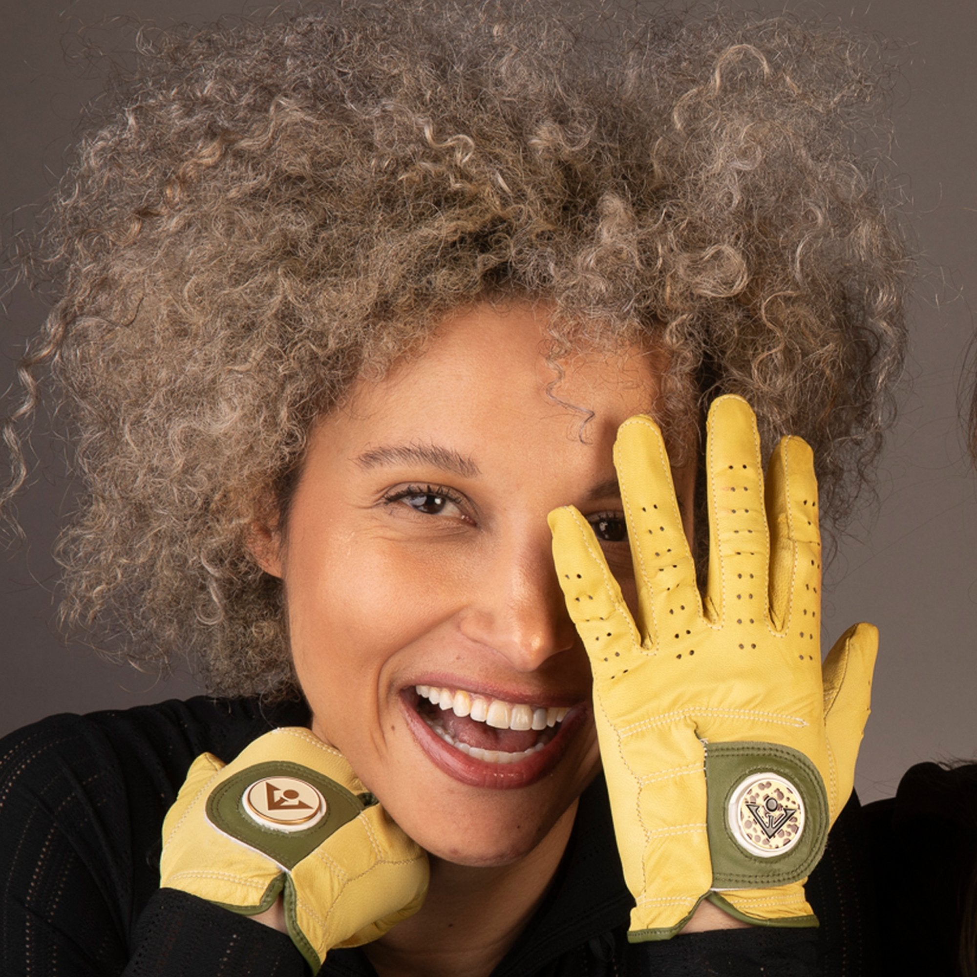 Radiant woman with curly hair smiling broadly, wearing yellow leather golf gloves with green accents and distinctive circular emblems on the cuffs.