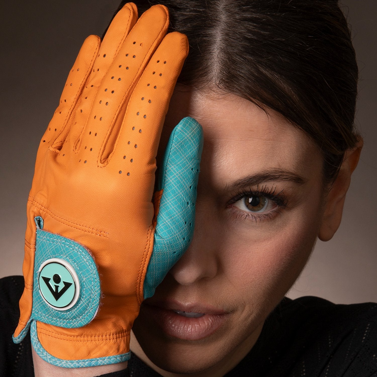 Woman holding a VivanTee Golf orange and blue accented golf glove in front of her eye staring into the camera up close.