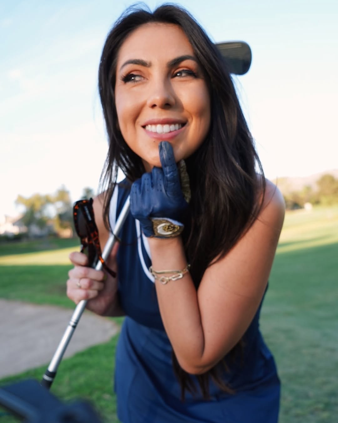 A close-up of a smiling woman golfer in a navy-blue golf dress, playfully touching her chin with a blue designer golf glove featuring a luxury gold emblem. She holds sunglasses in her other hand while resting a golf club on her shoulder, embodying confidence and elegance on the course.