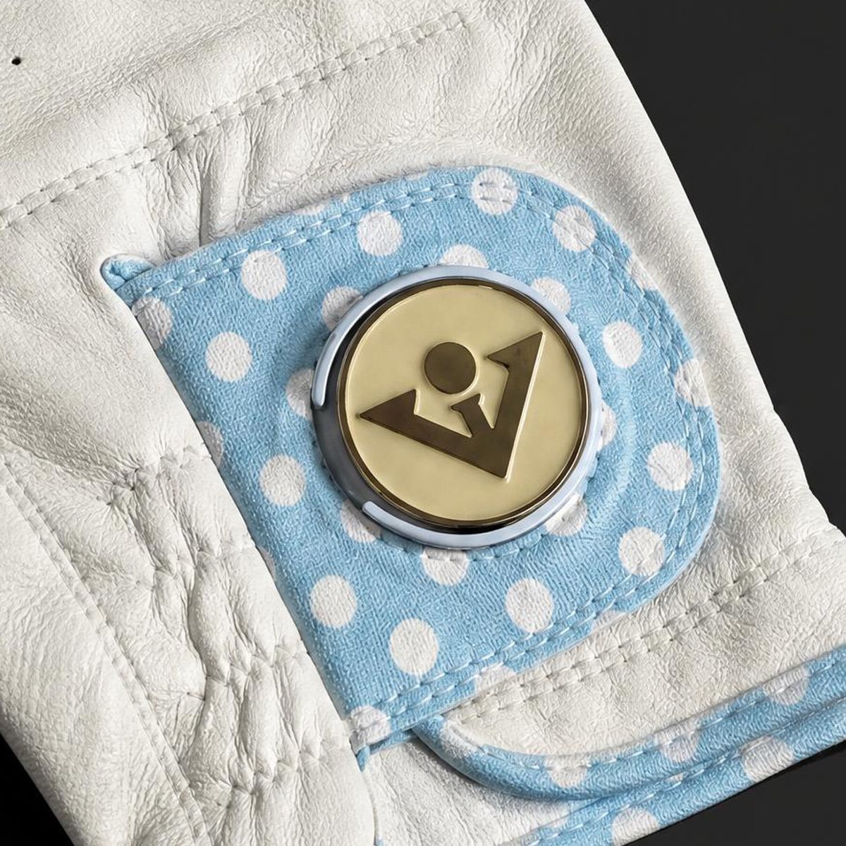 Close-up of a white leather golf glove with a light blue and white polka dot fabric patch pocket, featuring a round magnetic ball marker.  
