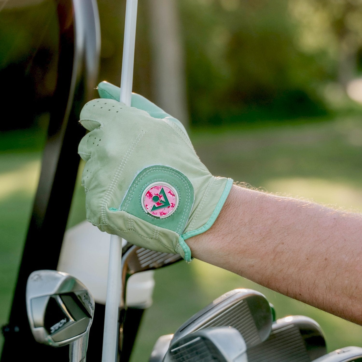 Close up of men's mint green golf glove with pink ball marker putting away a golf club in his bag.