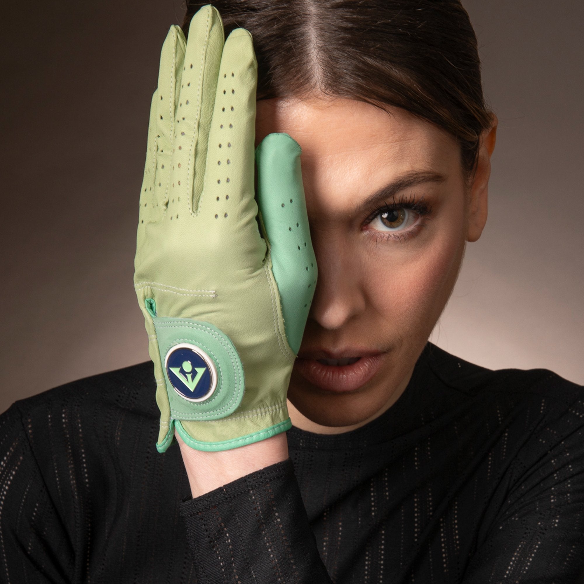  Intense gaze of a woman partially covering her face with a hand wearing a women's mint green golf glove, detailed with perforations and featuring a vibrant blue and green magnetic ball marker with VivanTee Logo..