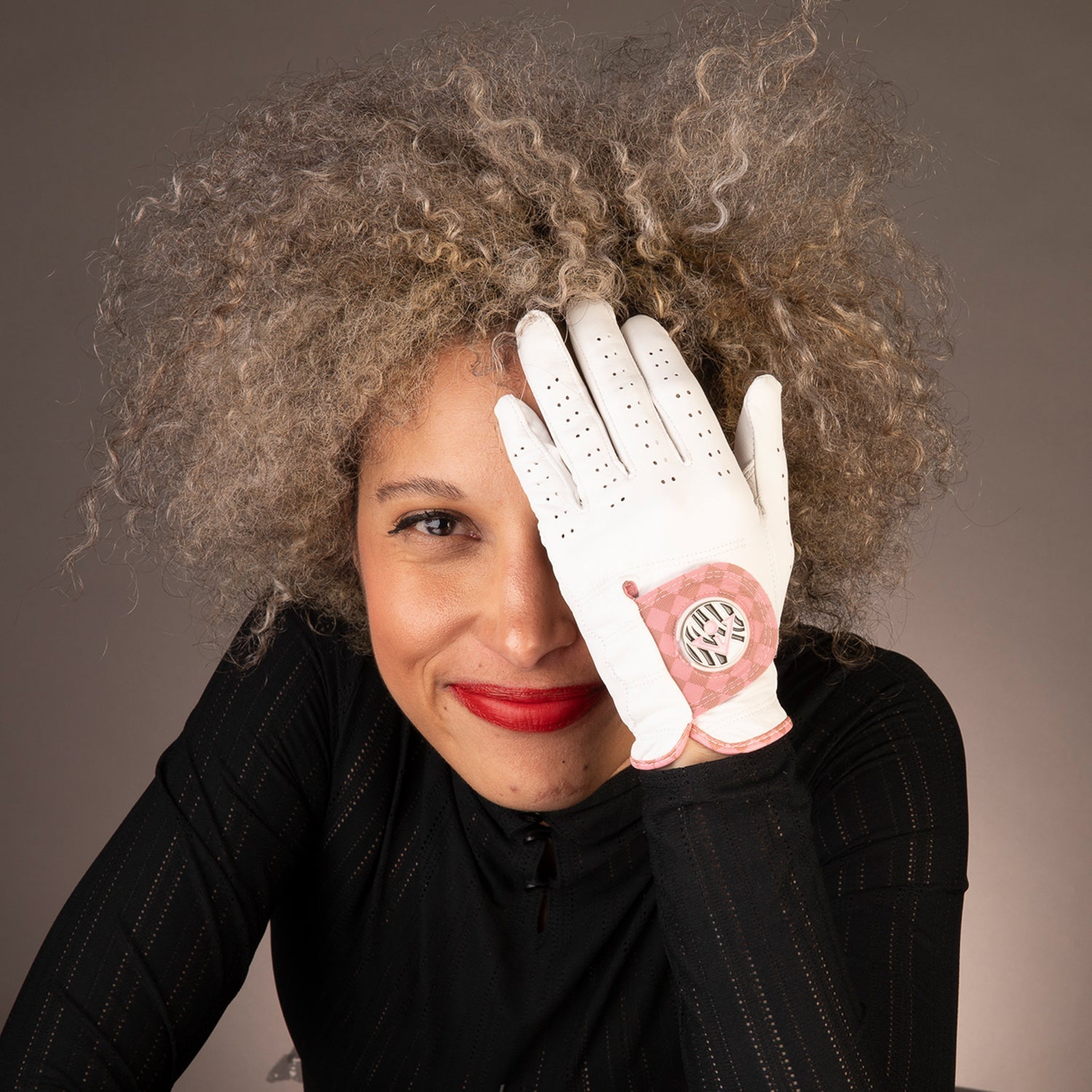 Woman with curly hair smiling at the camera, wearing a white leather golf glove with a pink and black emblem on the back, displayed on her raised hand.