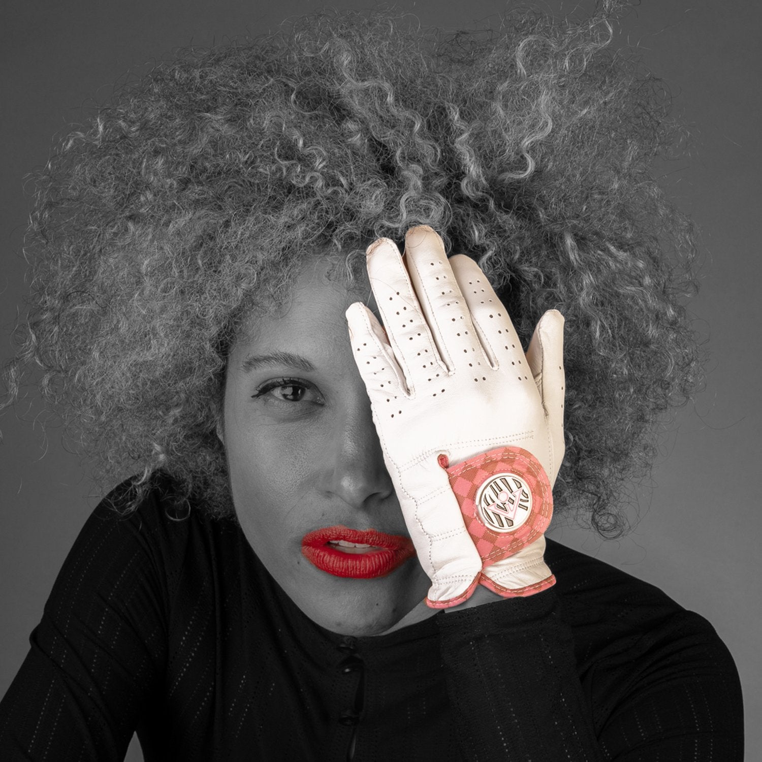Model wearing a white and pink golf glove with magnetic ball marker held up in front of her face in a monochromatic setting except the unique golf glove and her lipstick in red and pink.