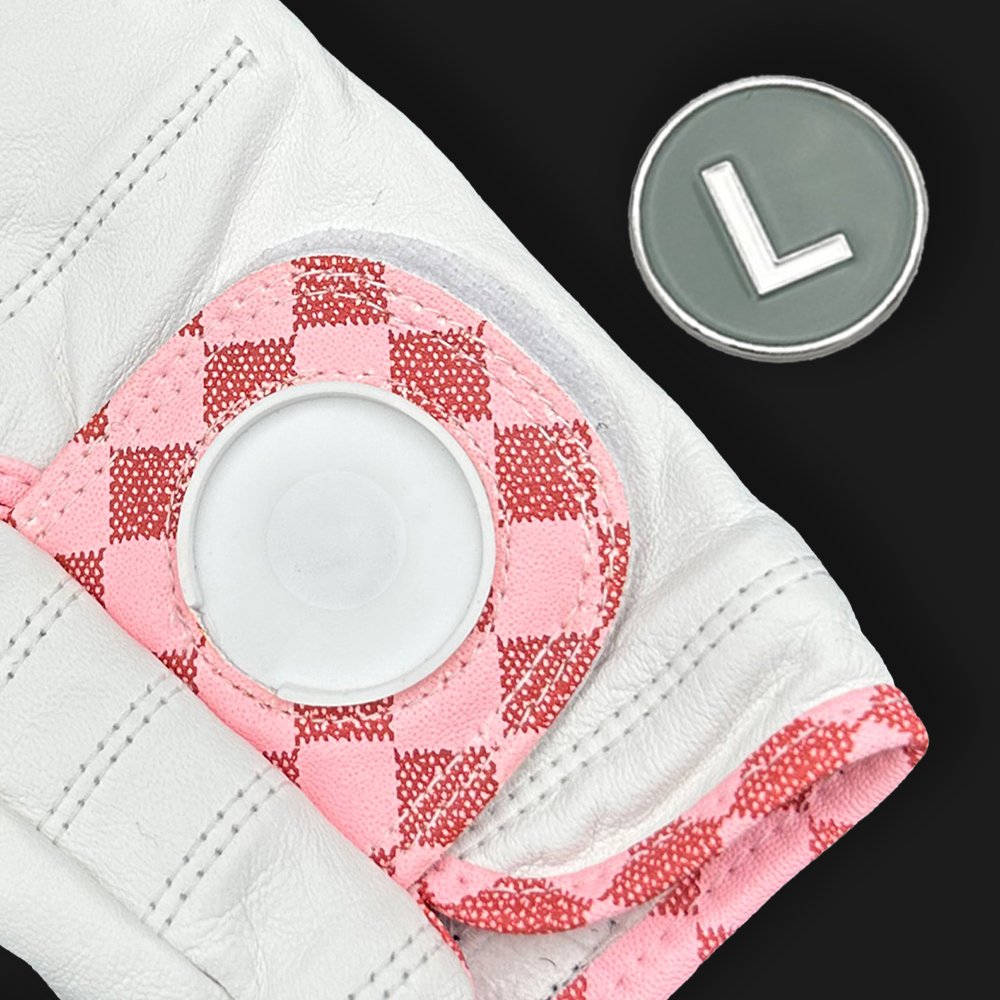 Close-up of a pink and red checkered golf glove with an L Train magnetic ball marker on a black background.