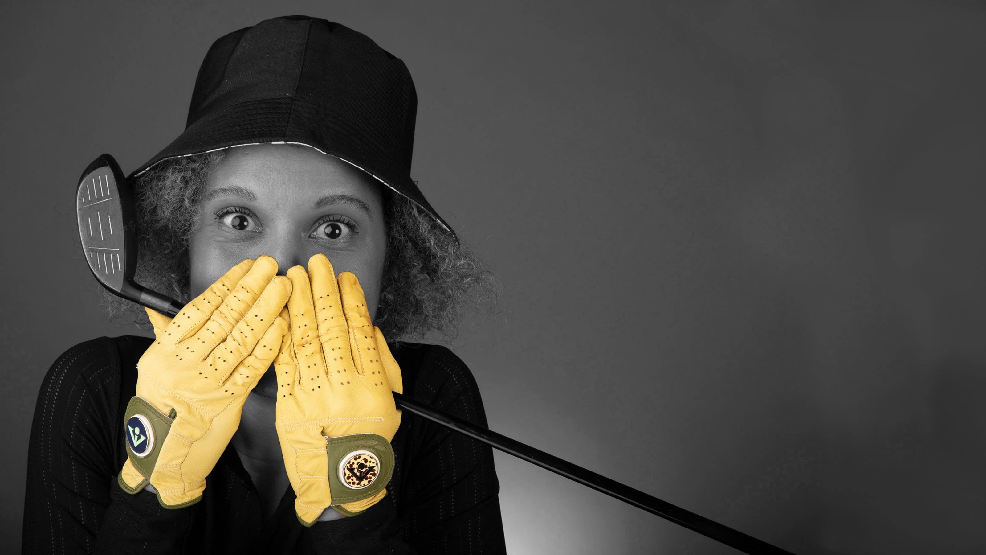 Woman posing with a golf club and bucket hat, wearing bright yellow VivanTee golf gloves with magnetic ball markers, against a monochromatic backdrop.