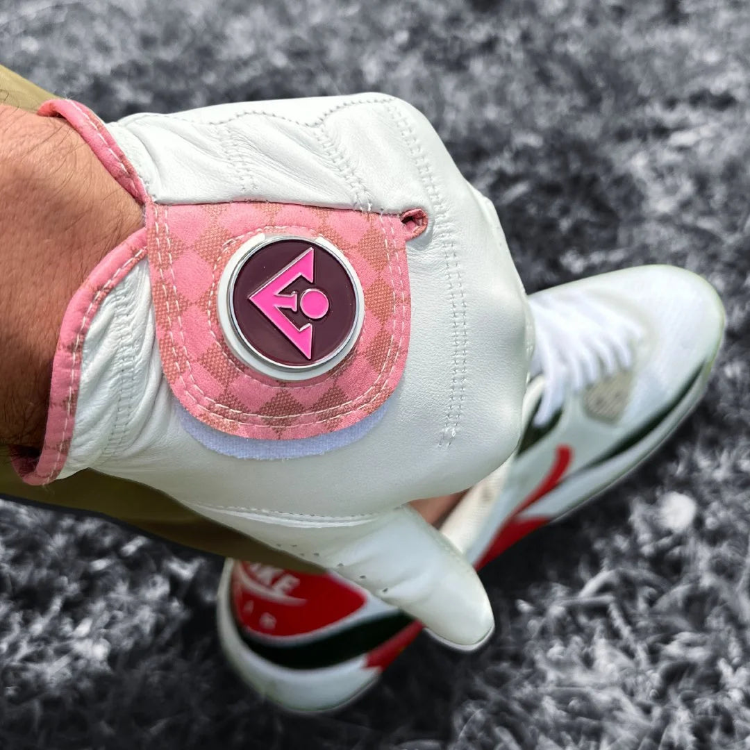 Golf glove with ball marker in white and pink with a black and white background, showing the red on the shoes in the background.