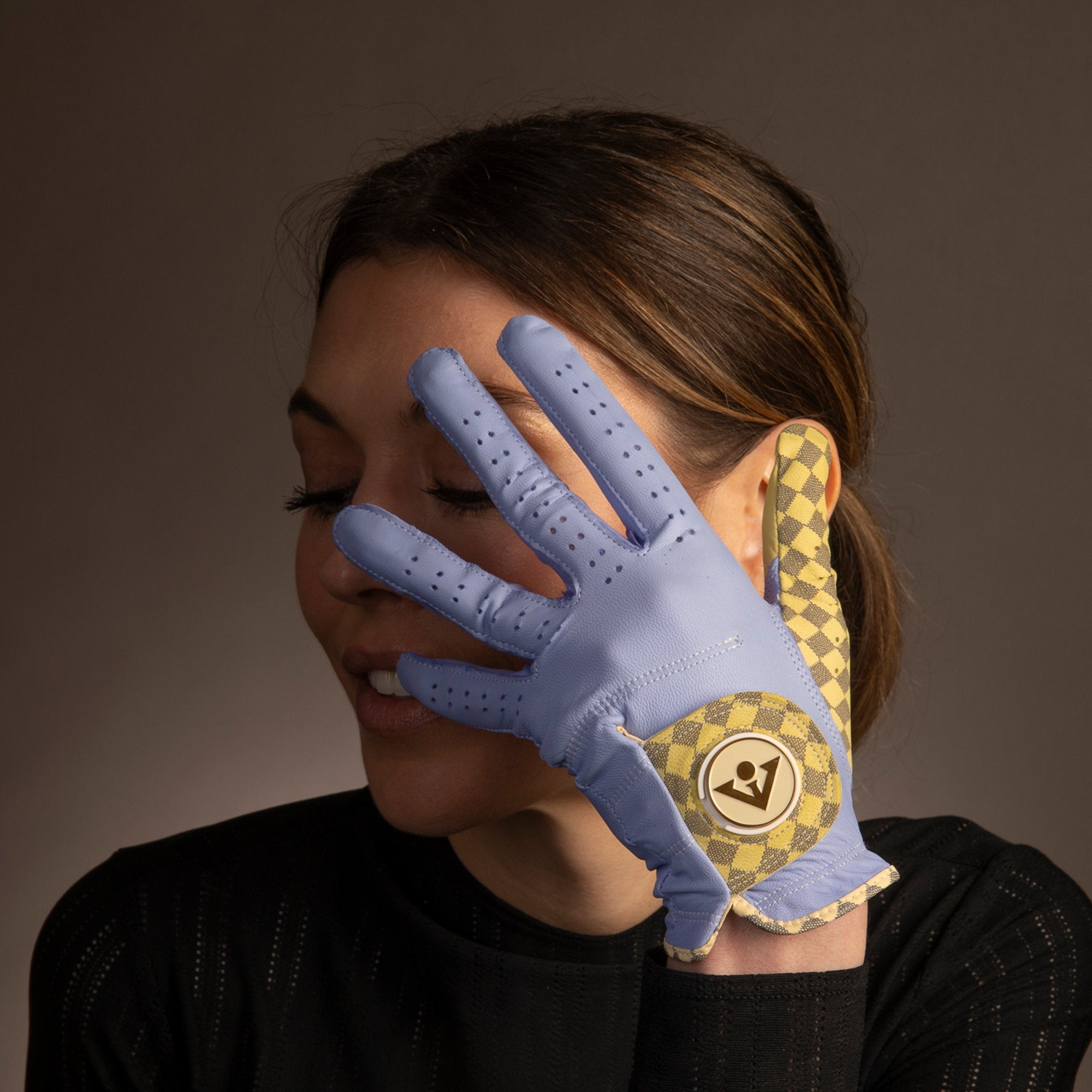 Alt Text: Side view of a woman with her eyes closed, gently touching her face with a hand adorned in a light purple leather golf glove featuring a yellow and gold checkered pattern and VivanTee Golf logo