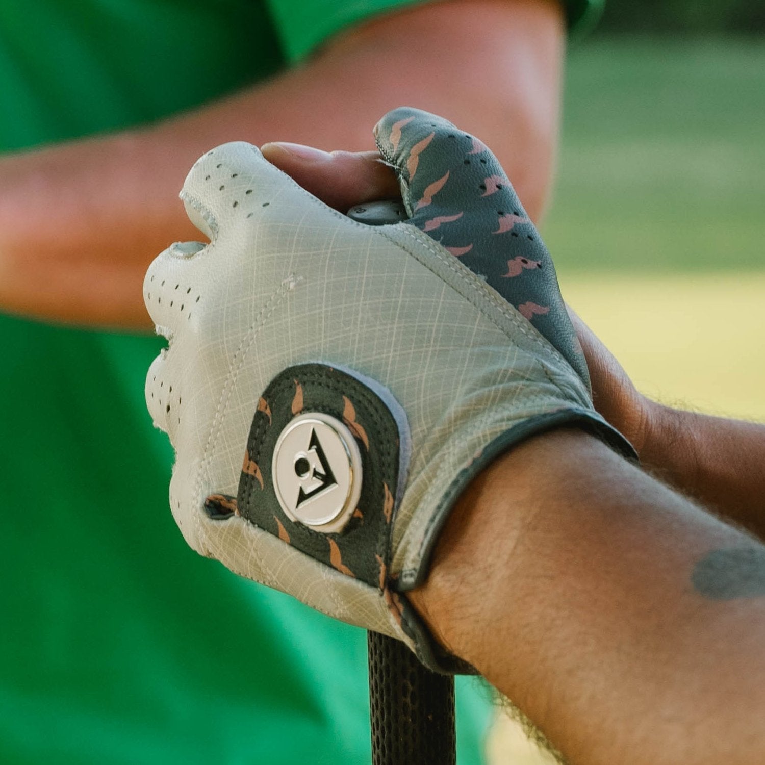 Men's charcoal golf glove with mustache pattern close up, holding the bottom of a golf club with other golfers in the background blurred, golf glove has a ball marker with VivanTee Logo in white and black.