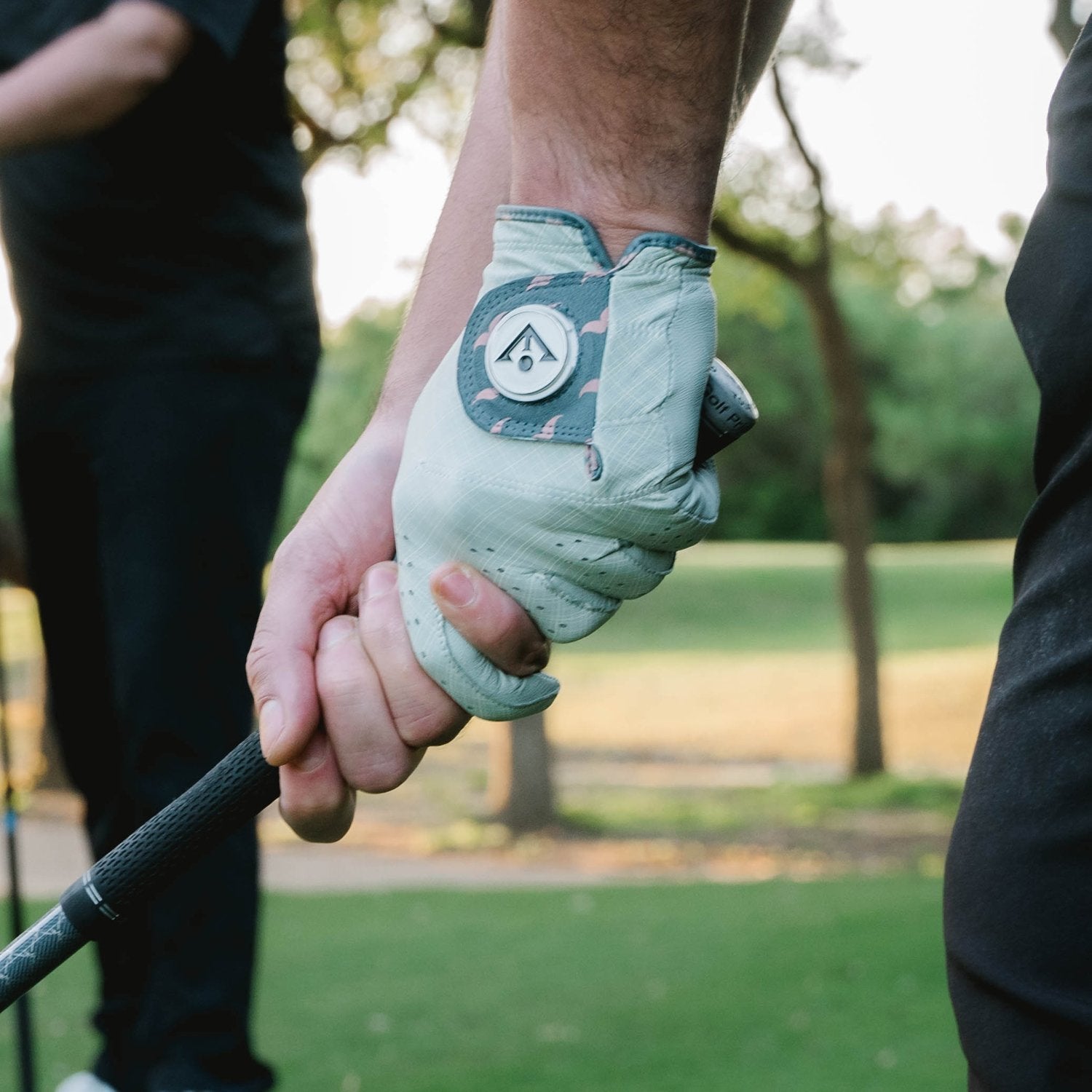 Close up image of men's gray golf glove with magnetic ball marker holding a golf club with an interlock grip on the teebox.