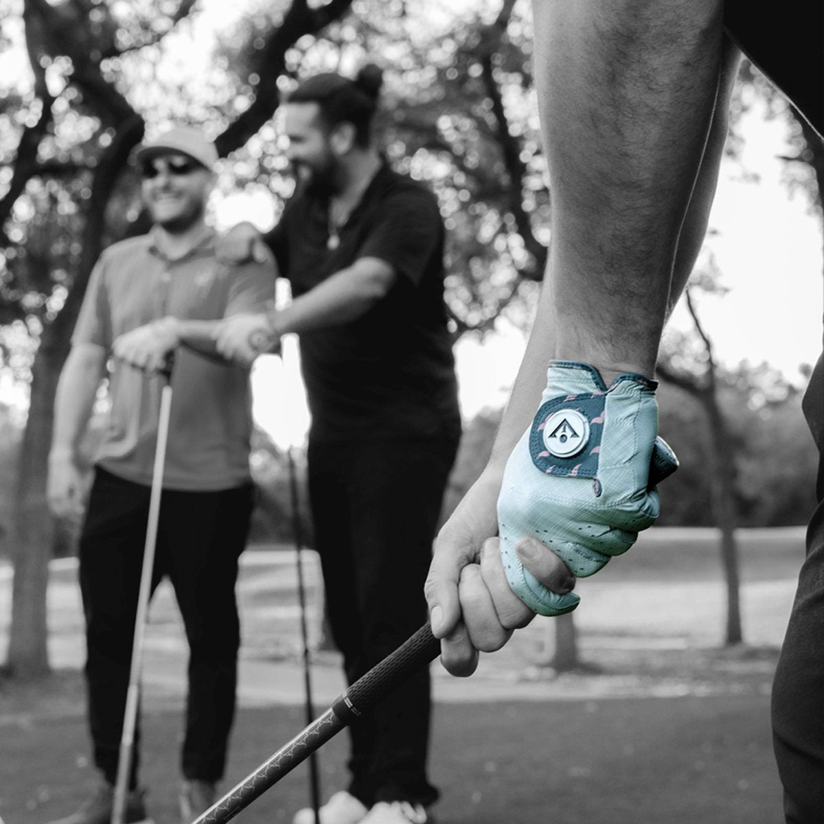 Person holding a golf club with a stylish golf glove, with two other people in the background laughing.  