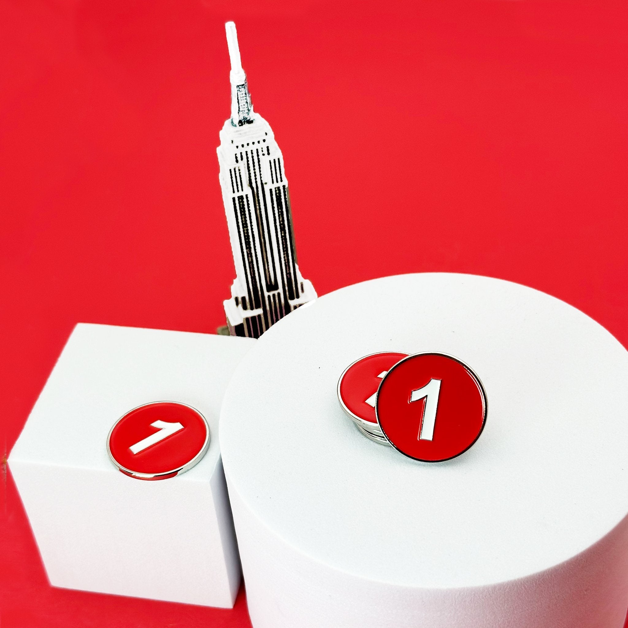 The NYC 1 Train Magnetic Ball Marker with a red backdrop to match the color, and the empire state building behind the product display.  Made for our golf gloves with ball markers.