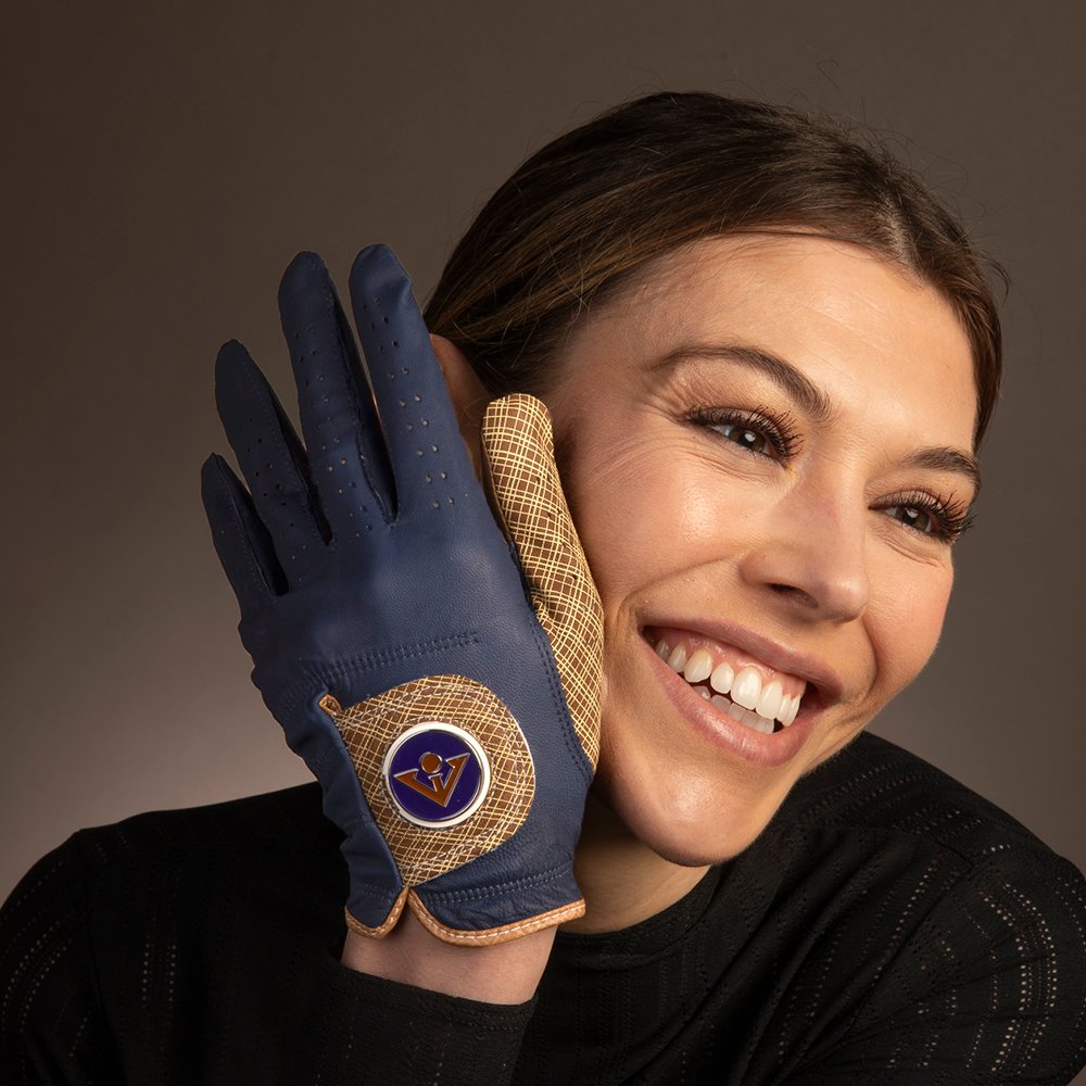 Navy Blue golf glove with brown accents being held up next to a models smiling face as she looks off to the left.