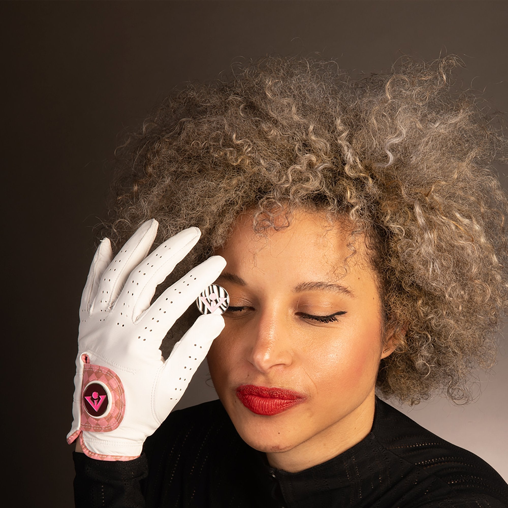 Model holding up a VivanTee ball marker in front of her right face while wearing a white and pink golf glove the SoHo Blush.